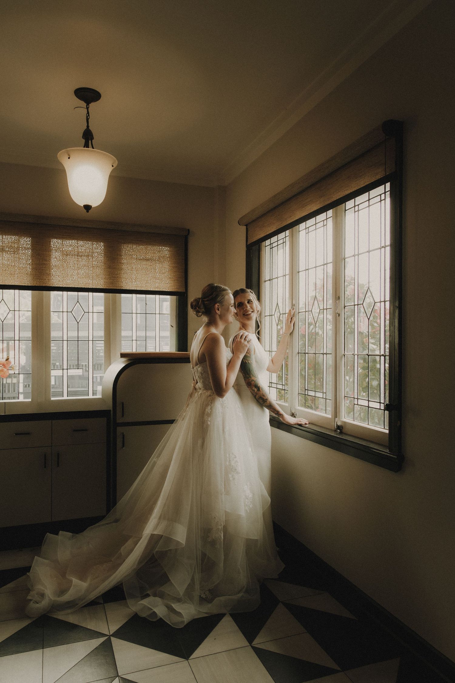 Two brides in white wedding gowns stand together by leaded glass windows in a warmly lit vintage room.