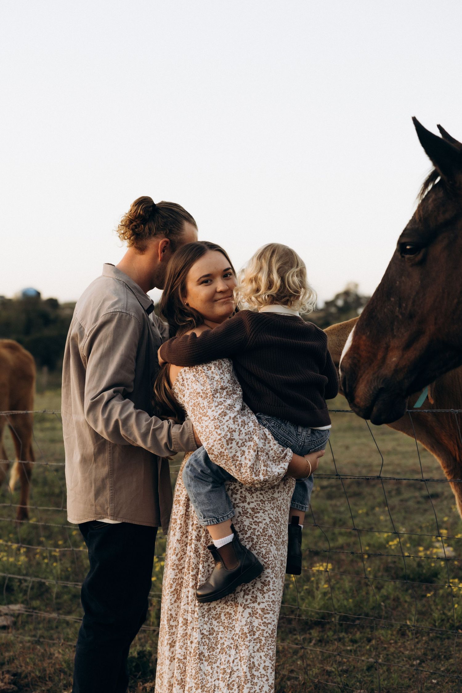 A couple holds their young child near a horse on a farm at golden hour, the woman smiling warmly in a floral dress.