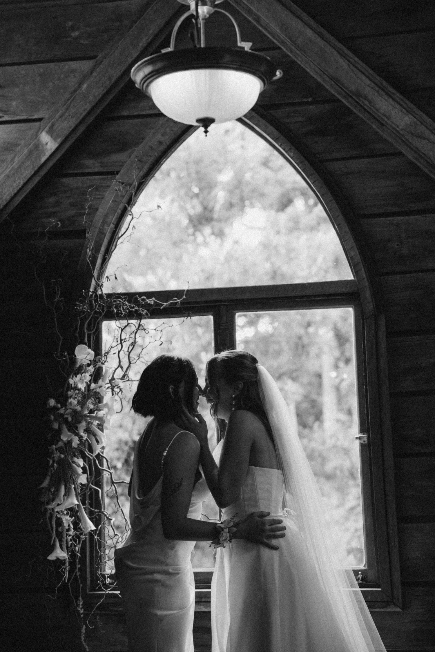 Two brides share an intimate moment in a rustic chapel with arched window in black and white wedding photo.