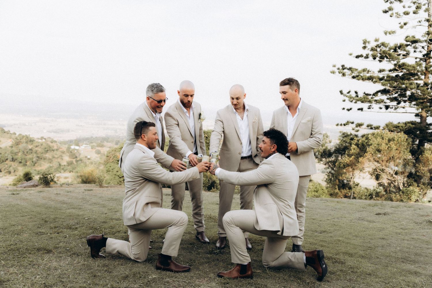 Groomsmen in beige suits kneel in circle formation outdoors.