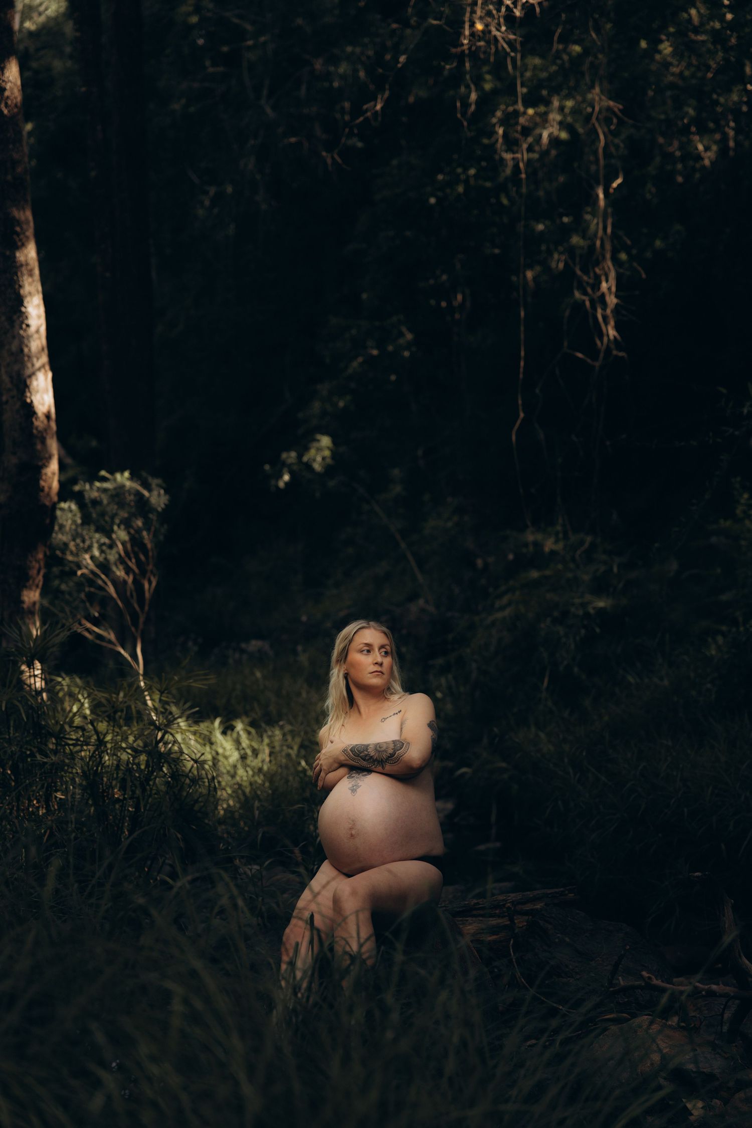Pregnant woman with tattoos posing in a lush dark forest, cradling her baby bump in a moody maternity photo.
