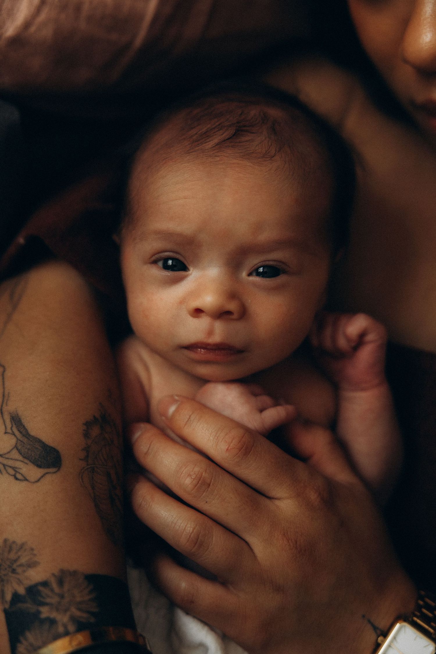 Newborn baby cradled in parents arms, with a tattoed forearm visible, in a warm intimate portrait.
