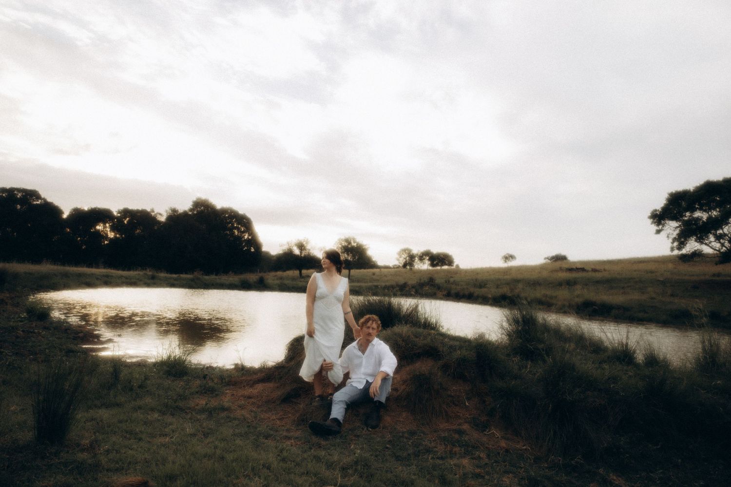 A couple sits together on a grassy hillside overlooking a scenic landscape at sunset.