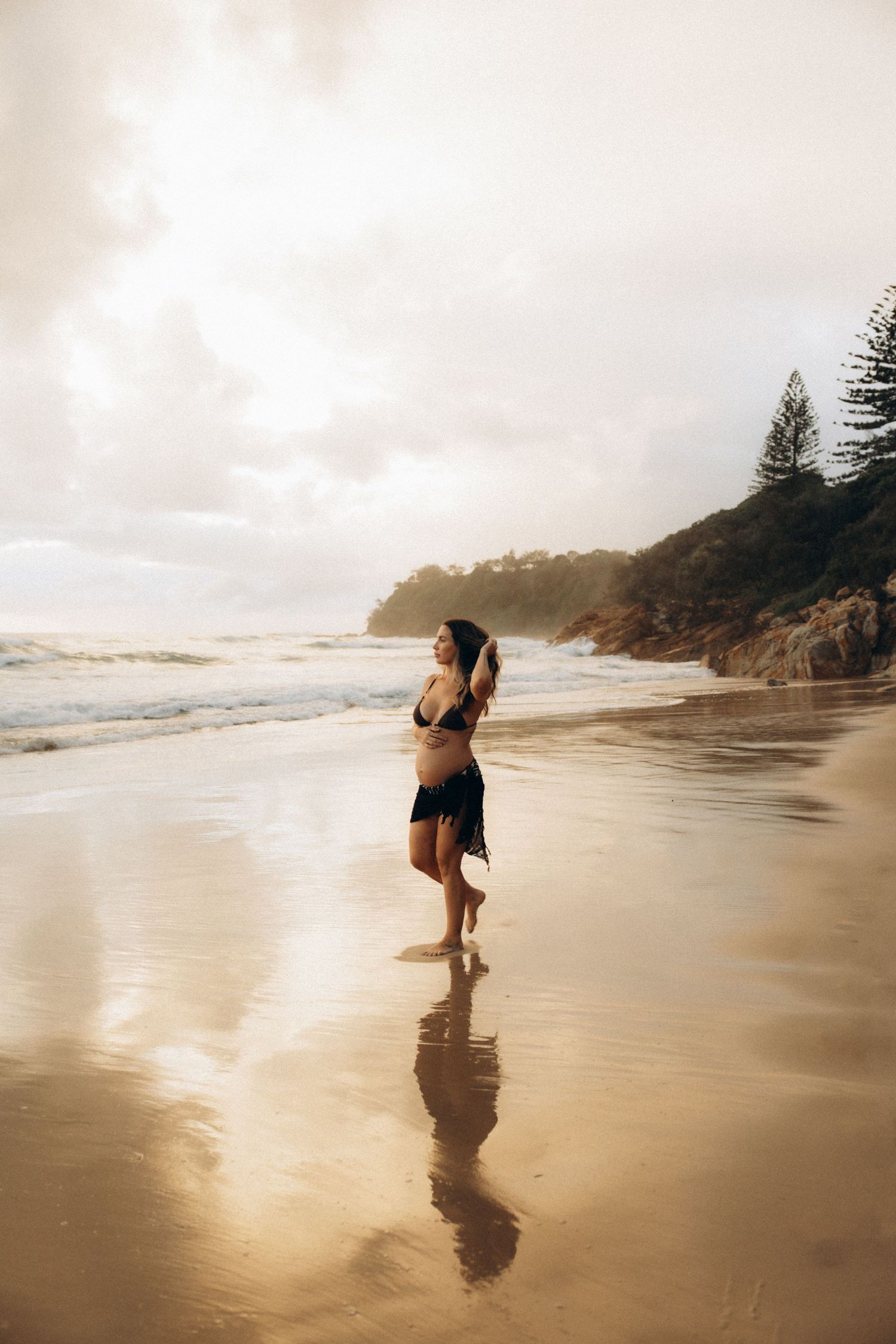 Silhouetted figure walking along a misty beach shoreline at sunset with cliffs in the background.