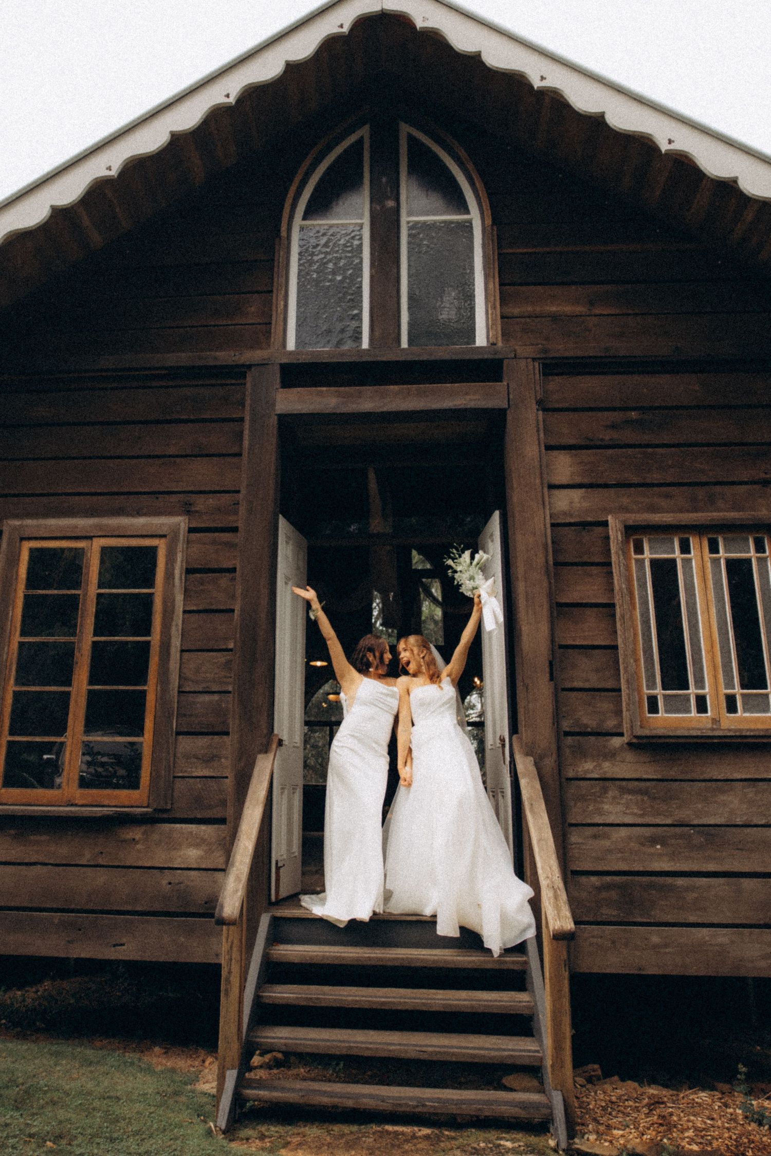 Two brides in white gowns raising bouquets at the entrance of a rustic wooden chapel on their wedding day.