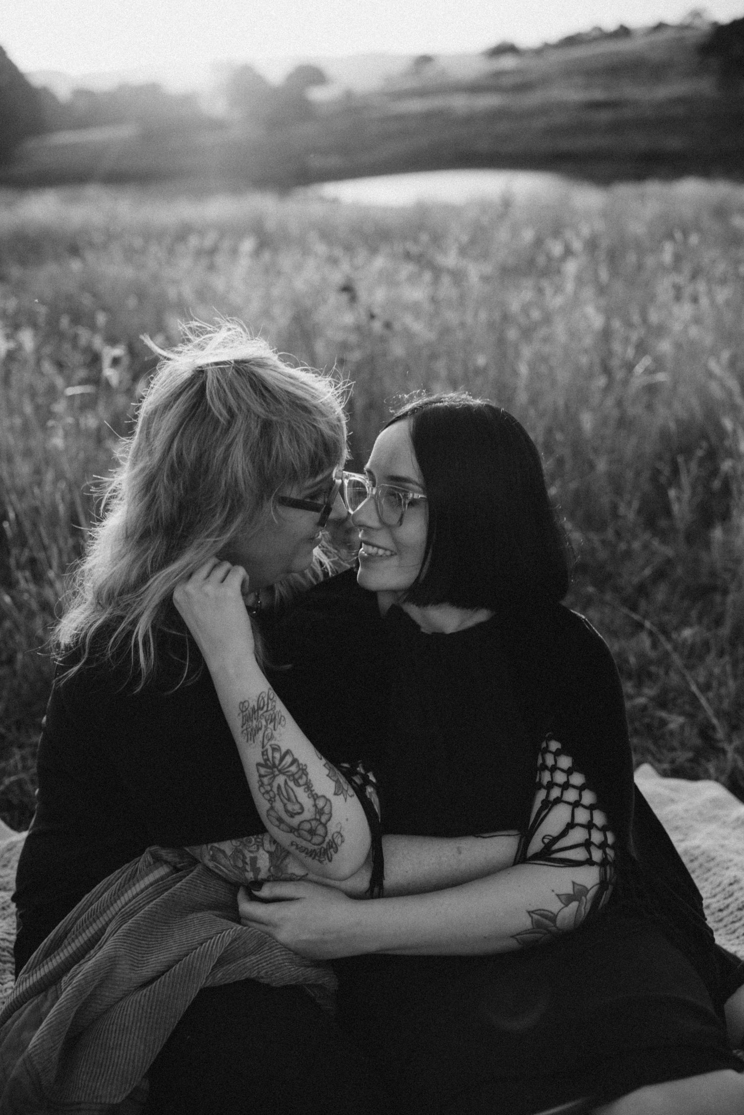 Black and white photo of two women embracing and smiling at each other in a field at sunset.