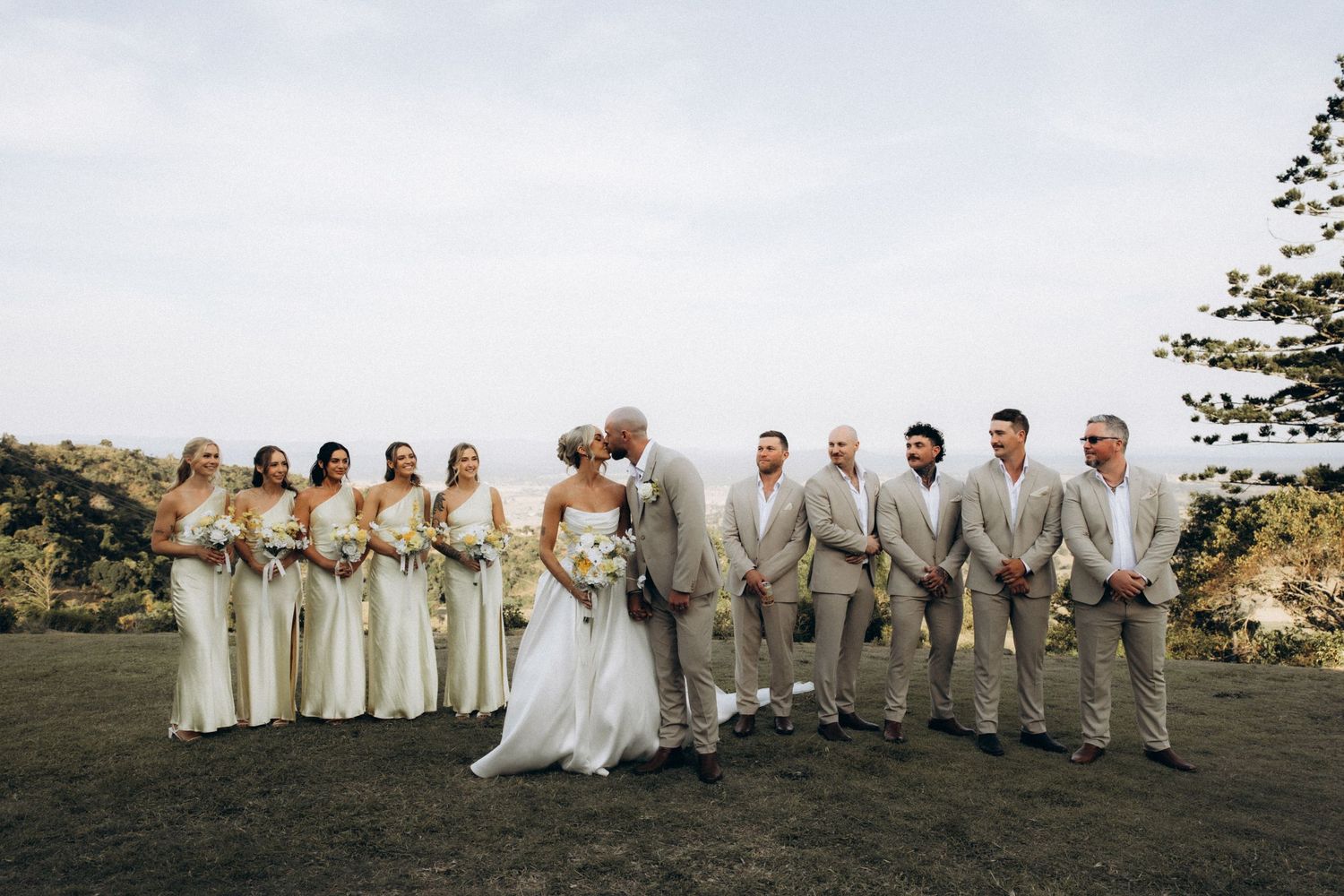 Full wedding party in cream and beige attire poses together on scenic overlook.