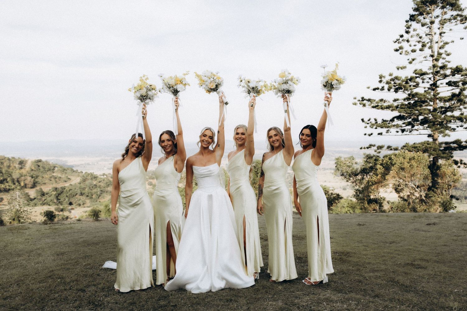 Wedding party in white dresses holding bouquets overhead while posing on a grassy hilltop.