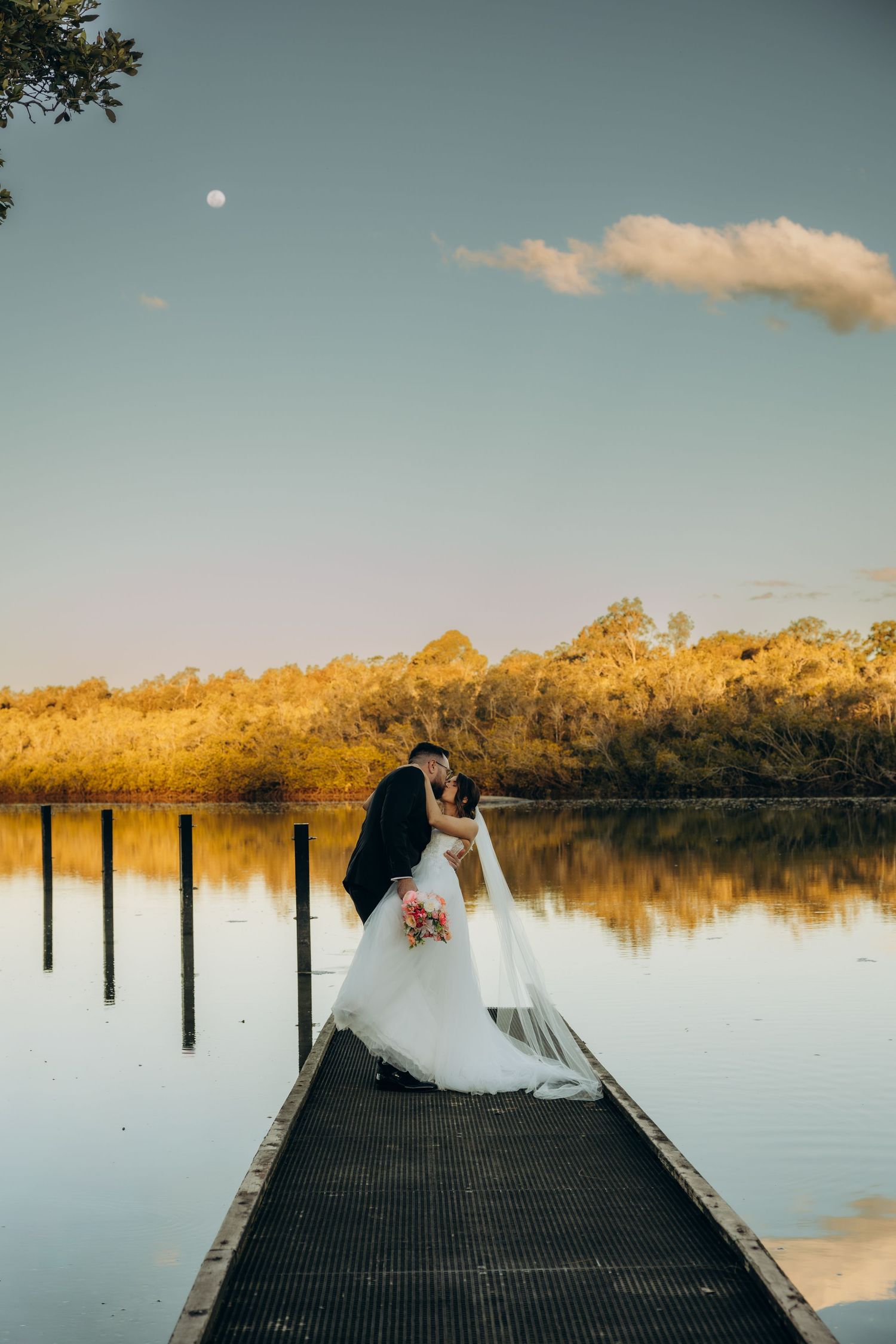 Bride and groom sharing a romantic kiss on a wooden dock overlooking a calm lake at golden hour.