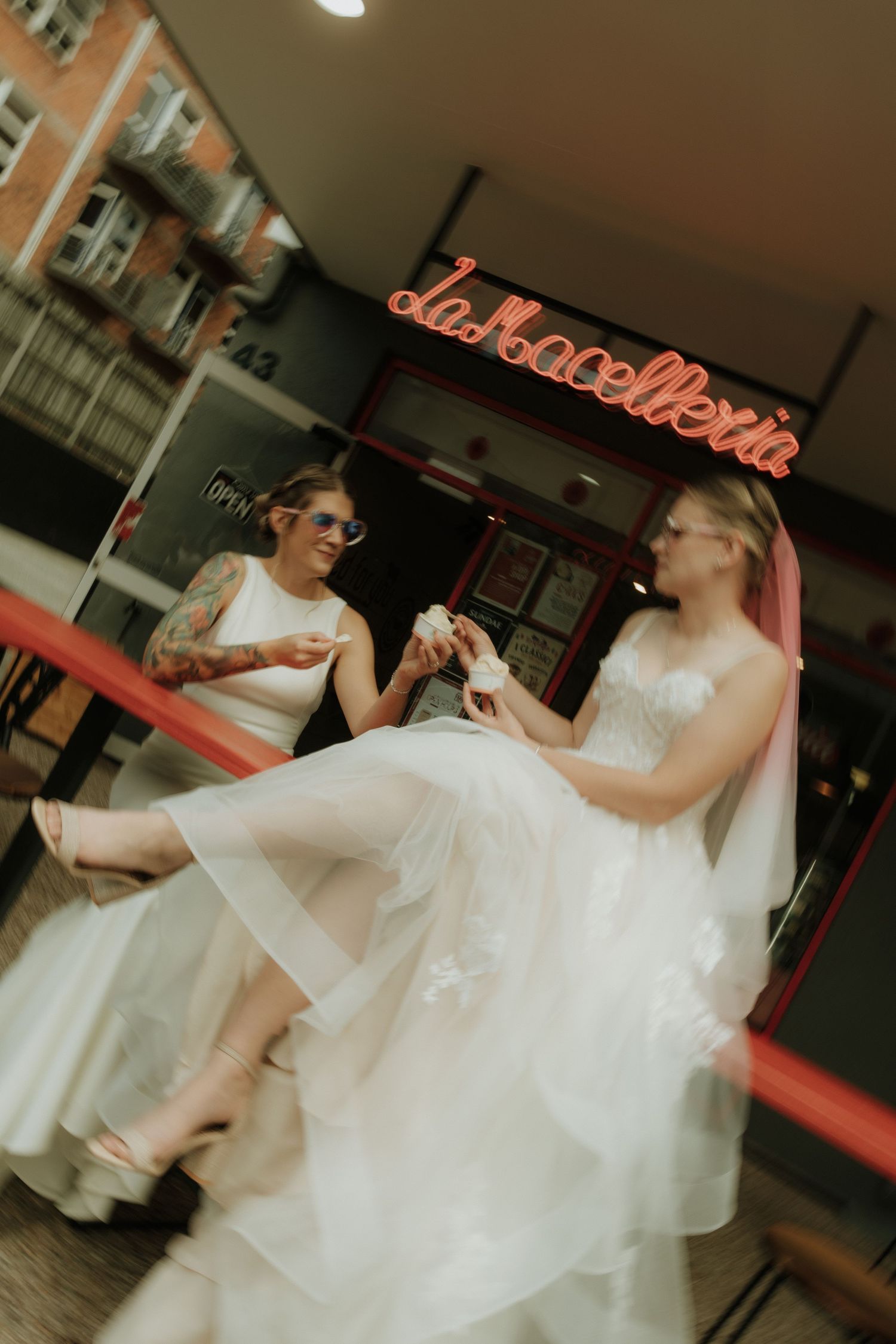 Two brides in white wedding dresses share a joyful moment outside La Macelleria restaurant, one with tattoos and sunglasses.