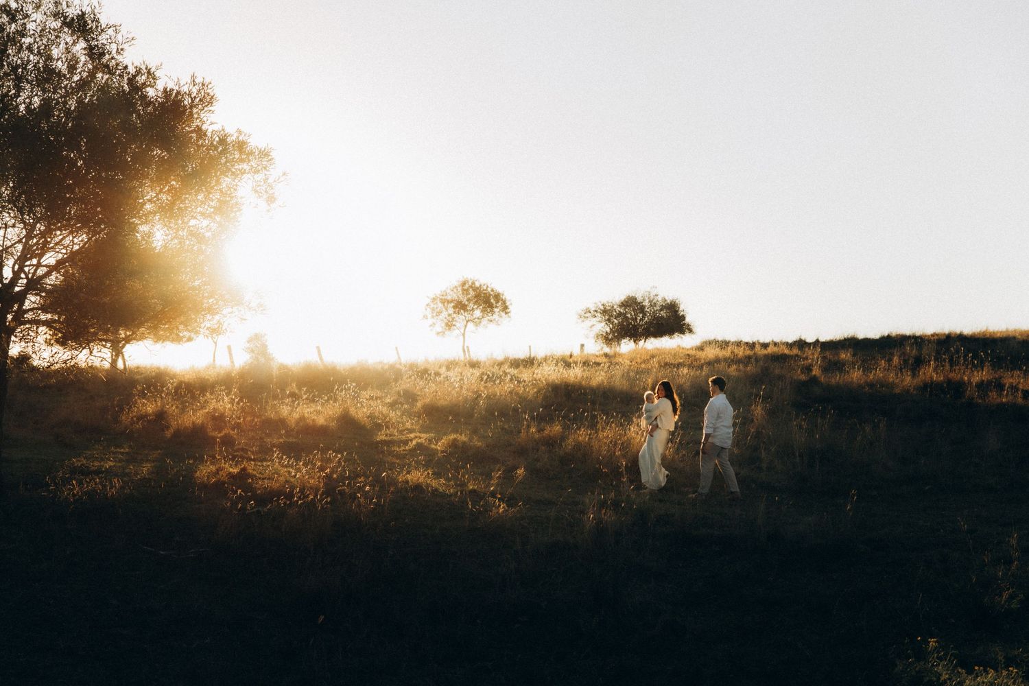 Couple walking hand in hand through golden sunlit fields at sunset with trees silhouetted against bright sky.