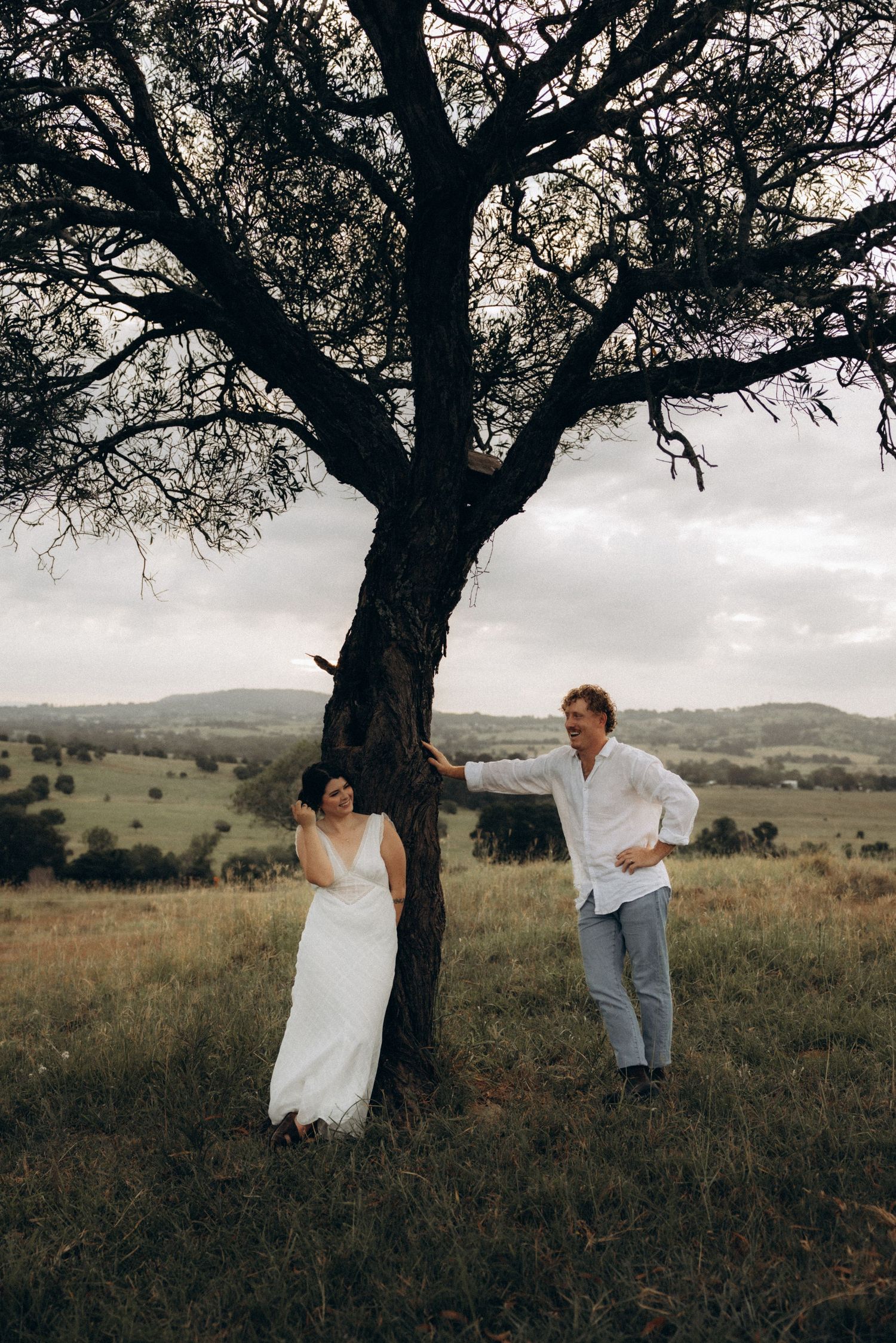 A playful couple poses under a large tree in a rustic field with rolling hills in the background.