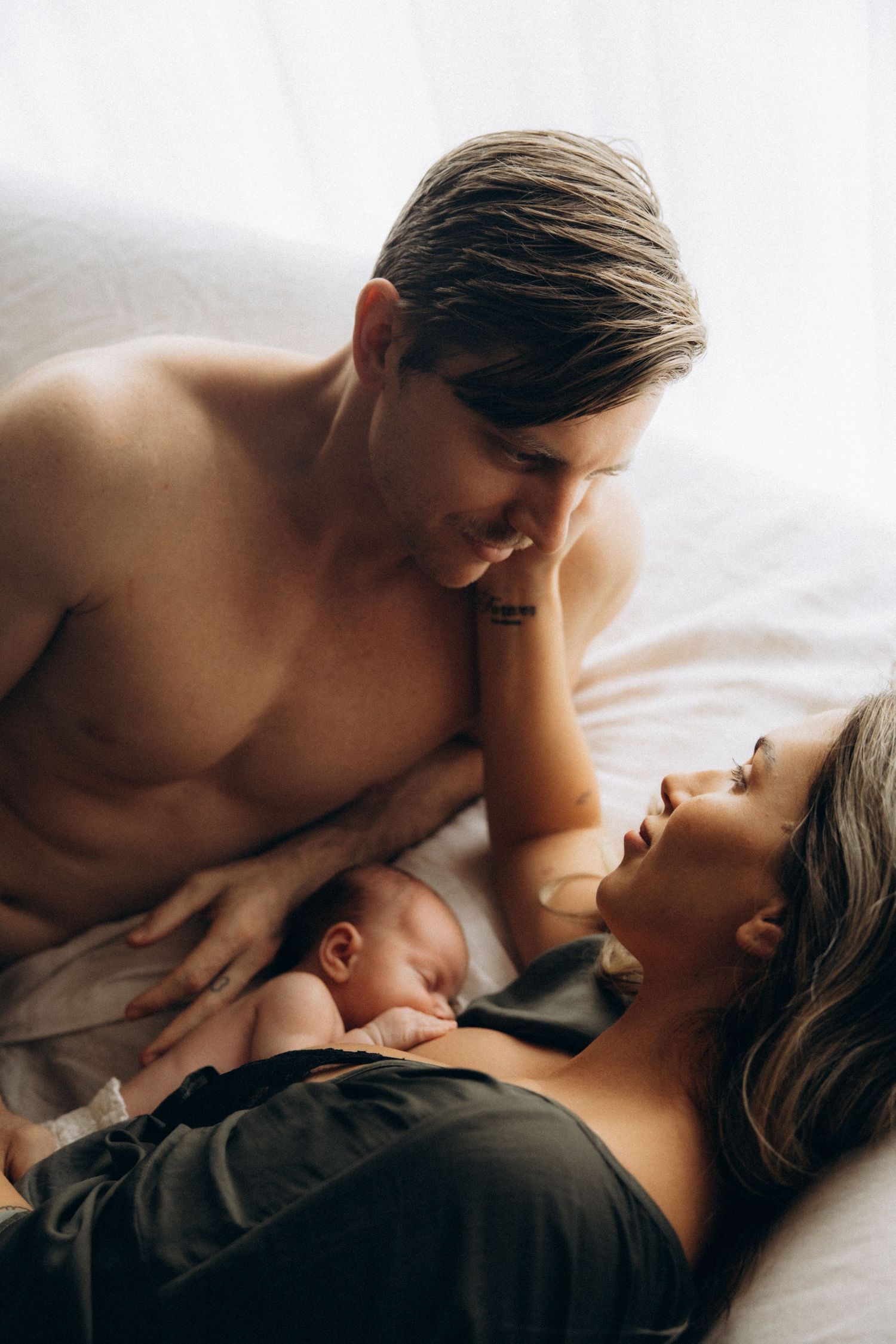 Intimate family moment with newborn baby sleeping peacefully between parents in bed.