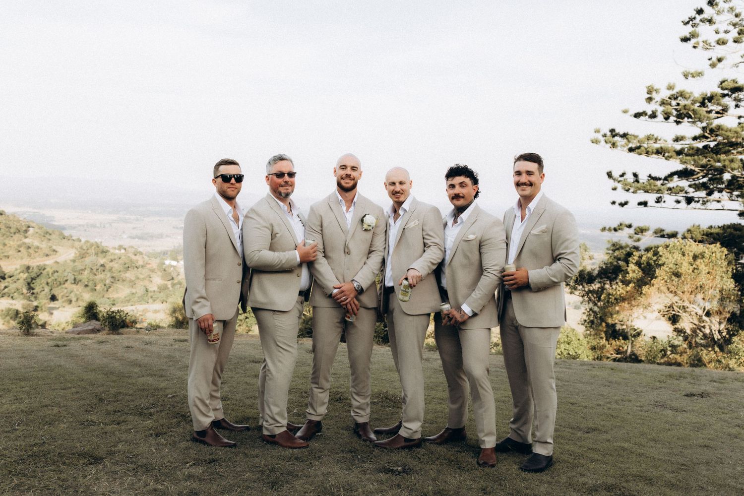 Groomsmen in light beige suits pose together on hilltop with scenic backdrop.