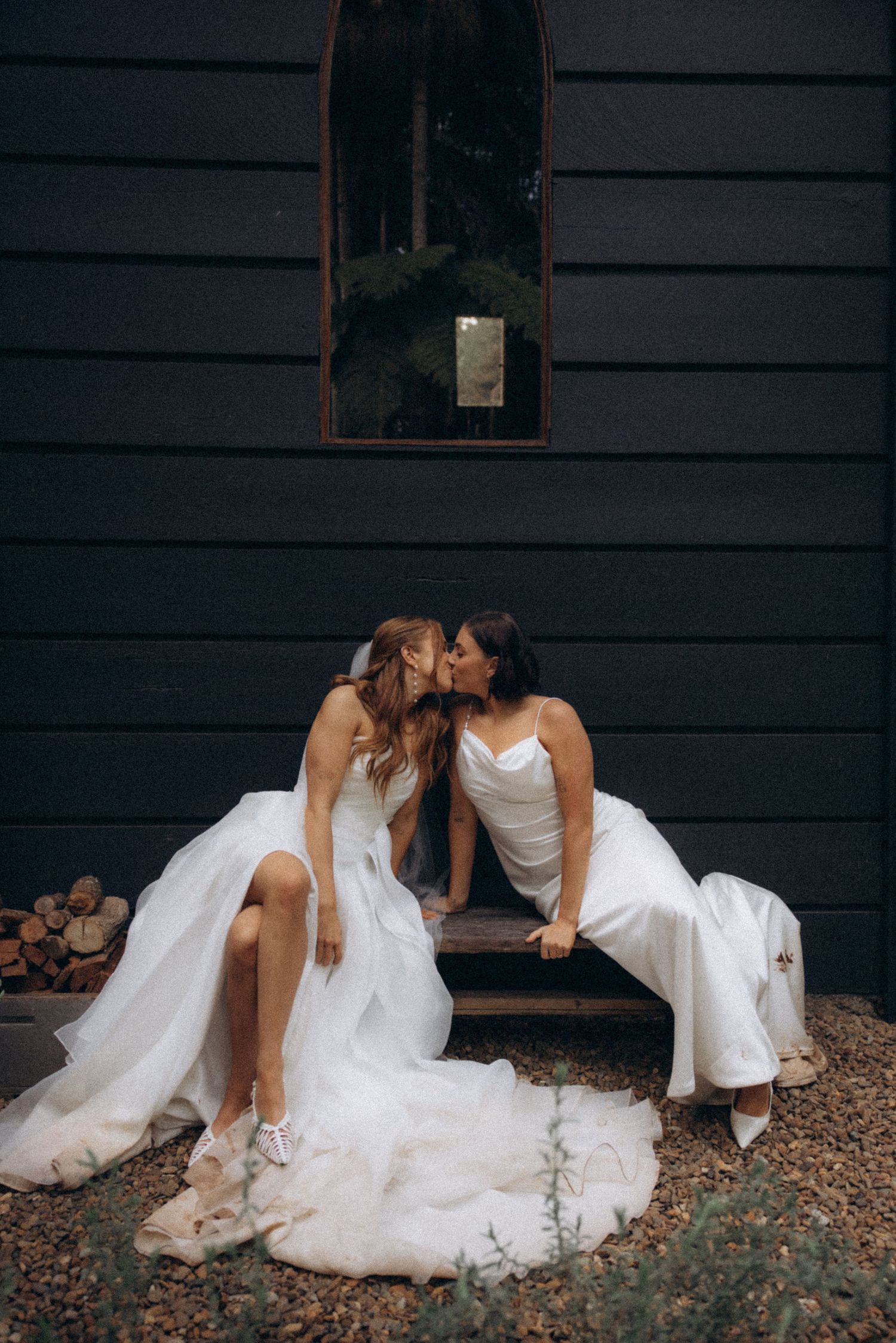 Two brides in white wedding gowns sharing a kiss on steps outside a dark barn venue.