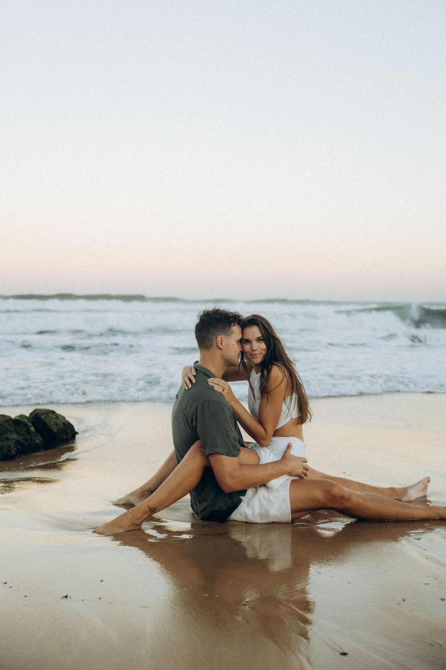 A couple sits embracing on a sandy beach at sunset, with ocean waves in the background.