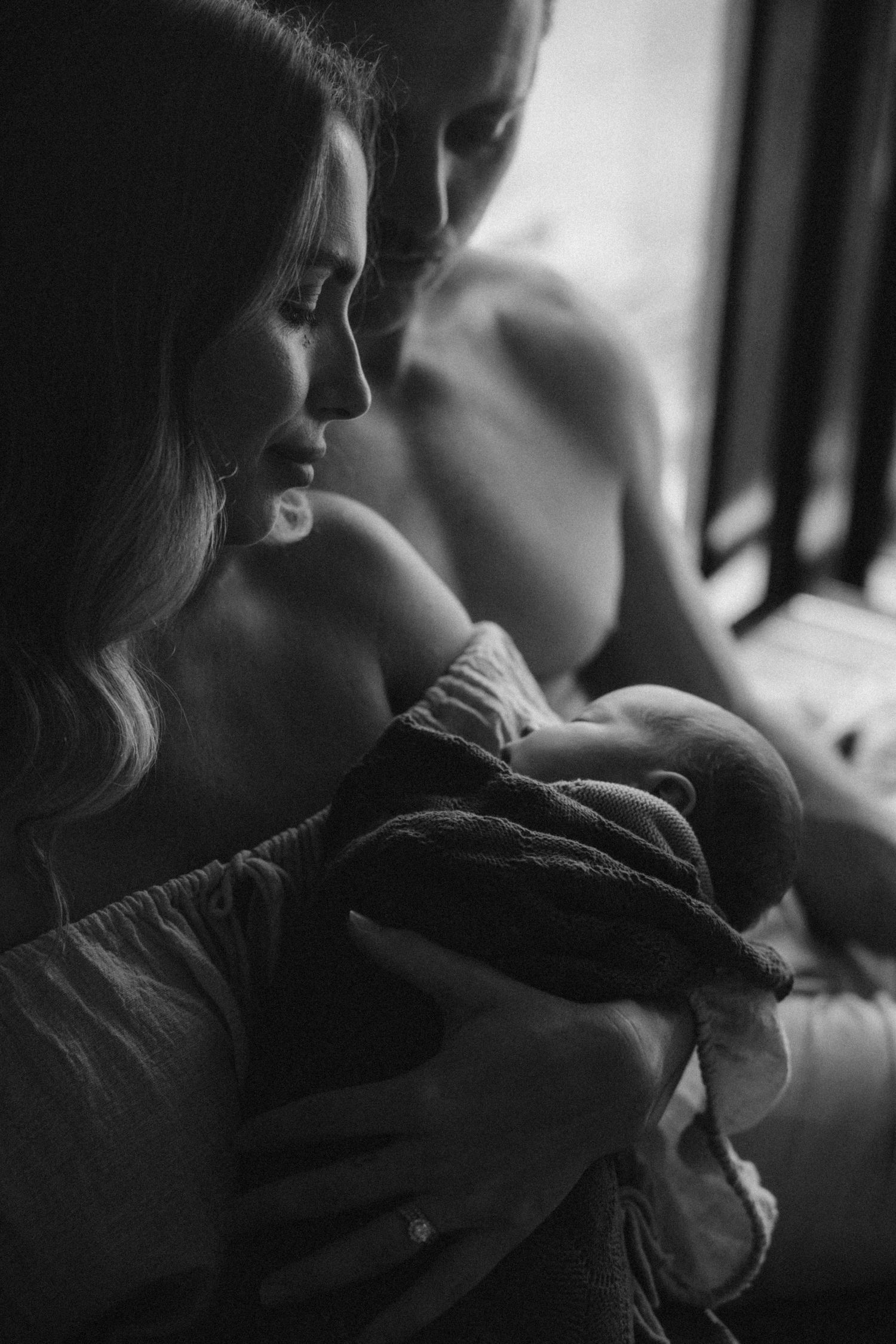 Intimate black and white portrait of parent holding sleeping newborn by window.