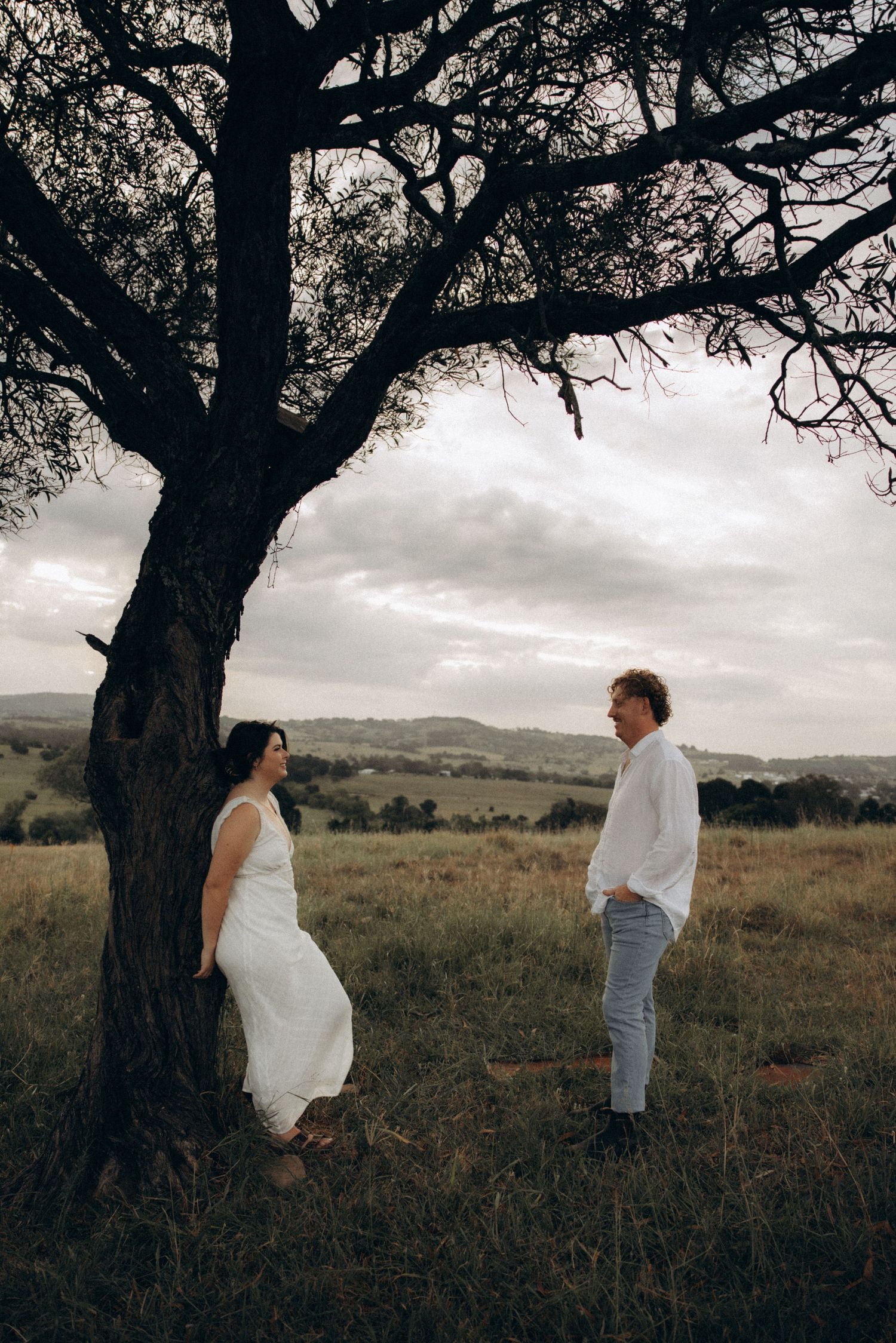 A couple plays hide and seek around a large tree in an open field.