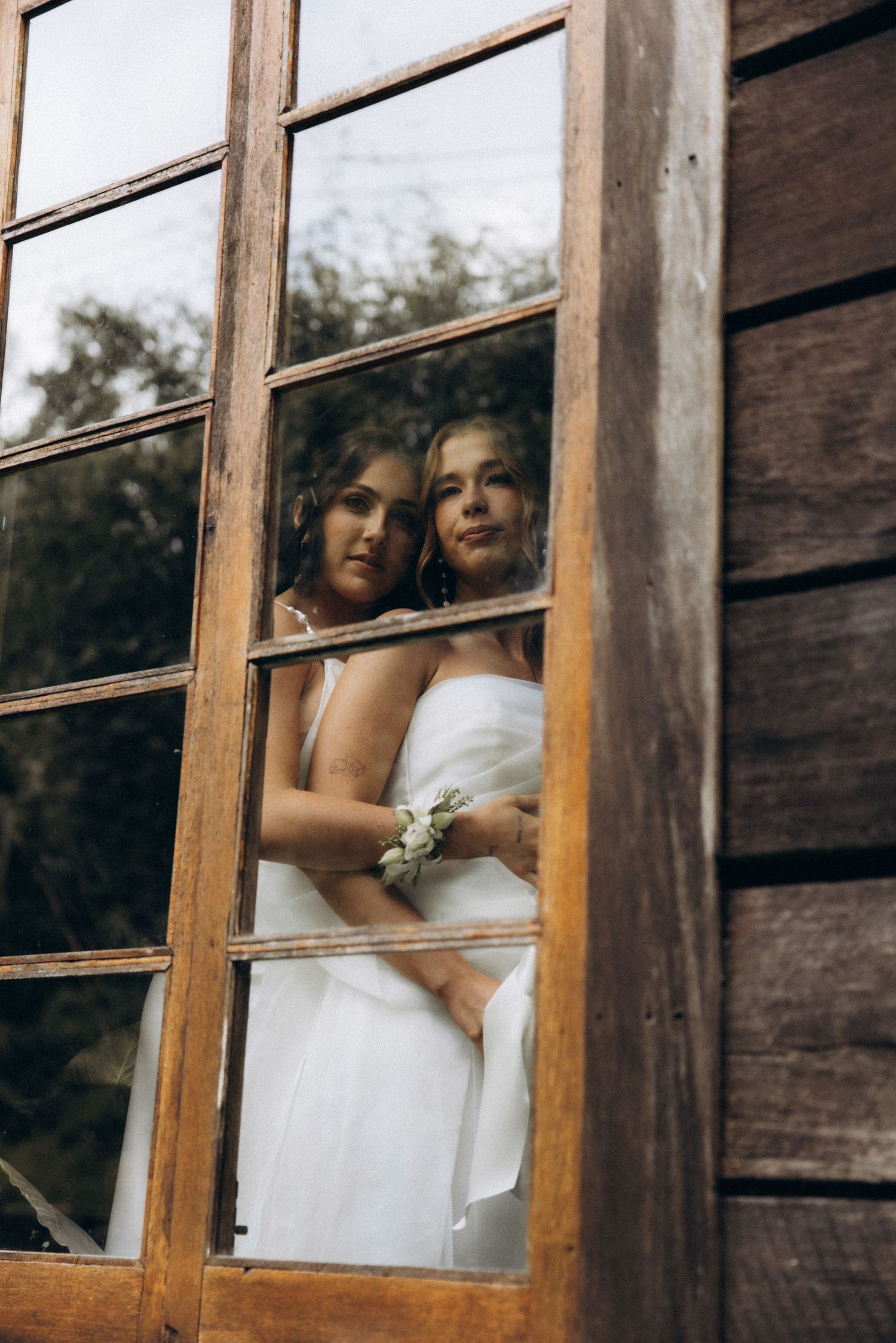 Two brides in white dresses embrace while looking through a rustic wooden window, captured in a romantic wedding portrait.