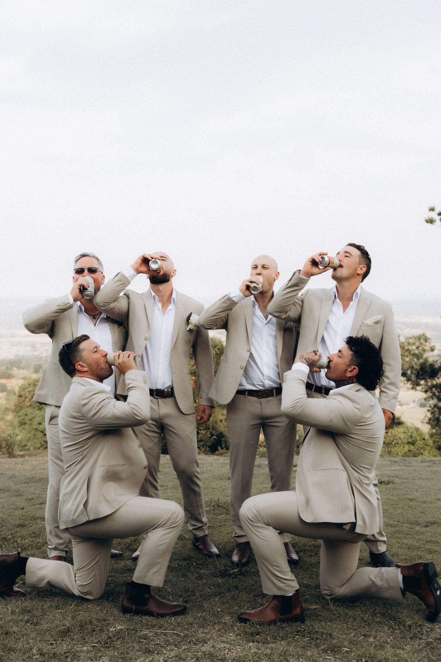 Groomsmen in light suits pose dramatically while pretending to drink.