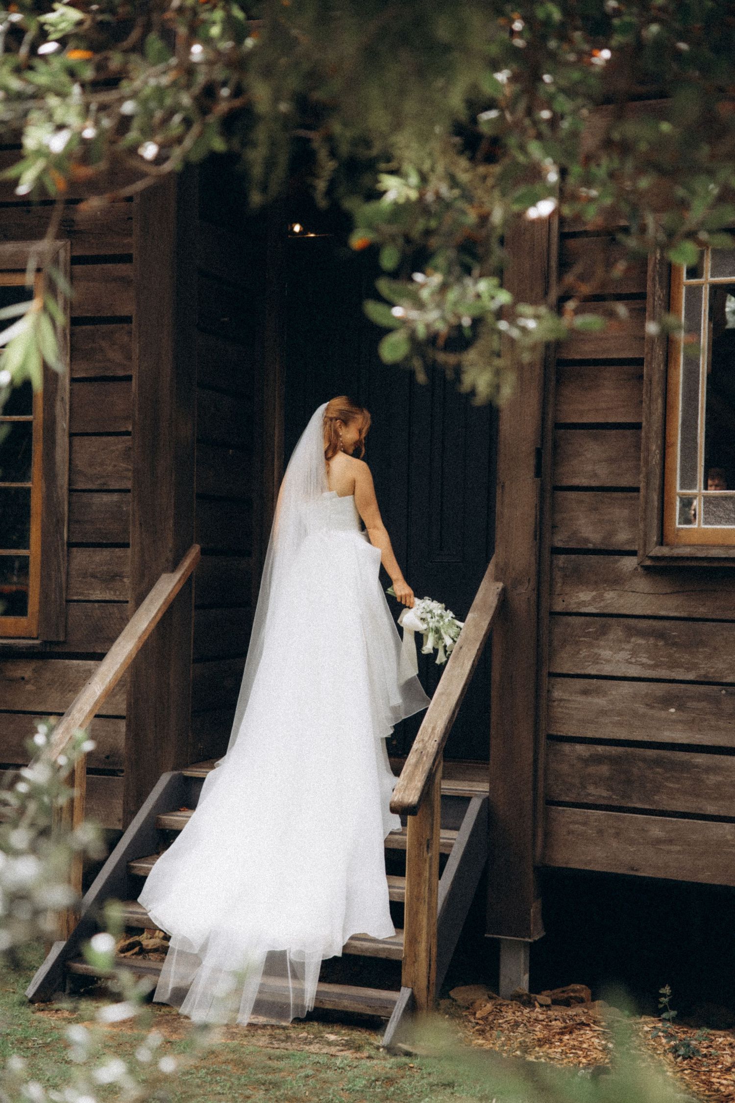 Bride in white gown and veil ascending wooden steps of rustic cabin, holding bouquet, surrounded by lush greenery.