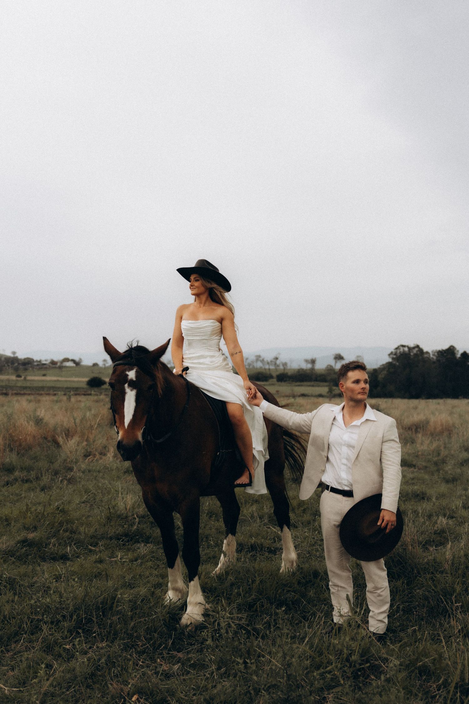 A couple poses with a beautiful dark draft horse against an overcast sky.