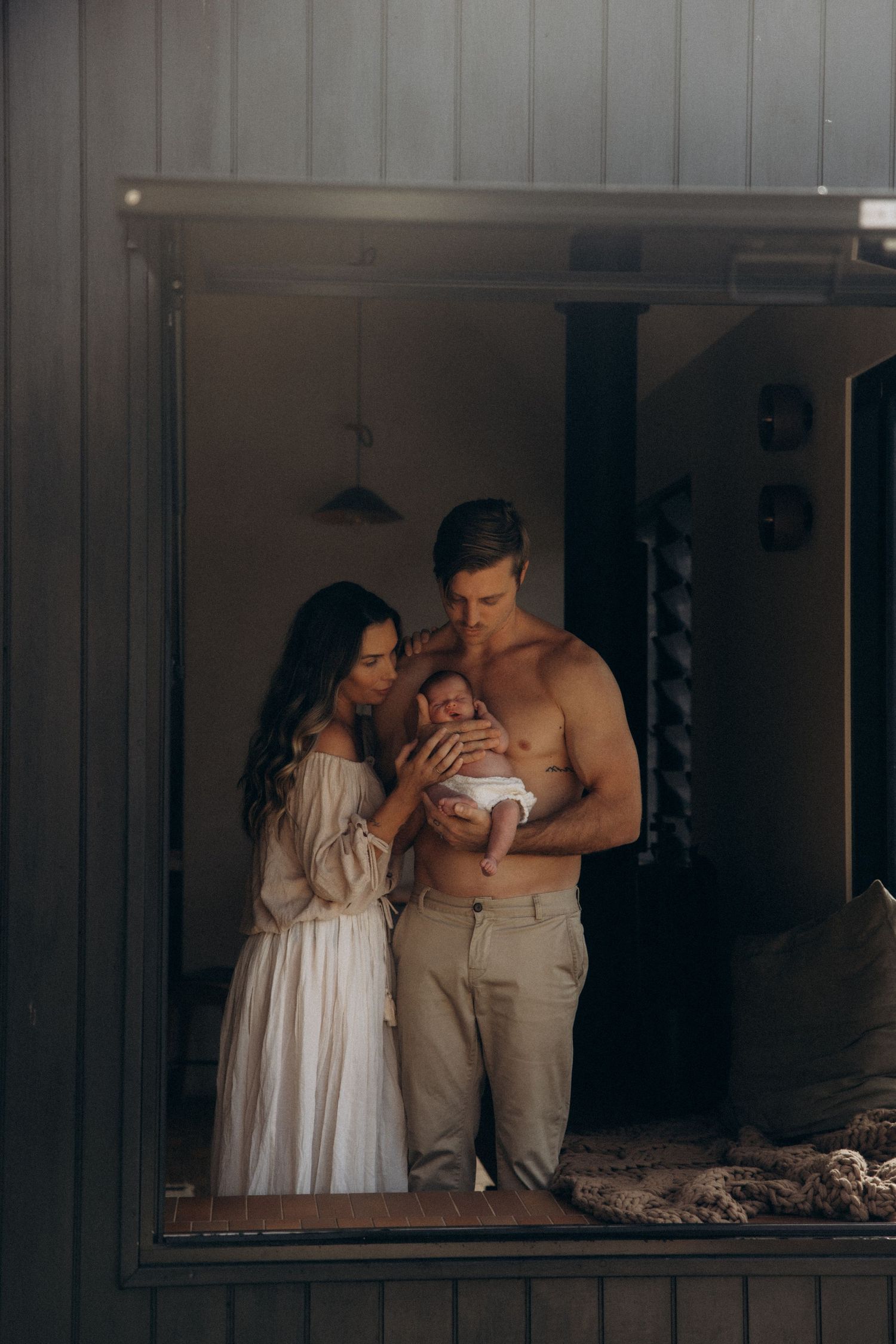 Family portrait reflection in mirror shows parents holding newborn in rustic setting.