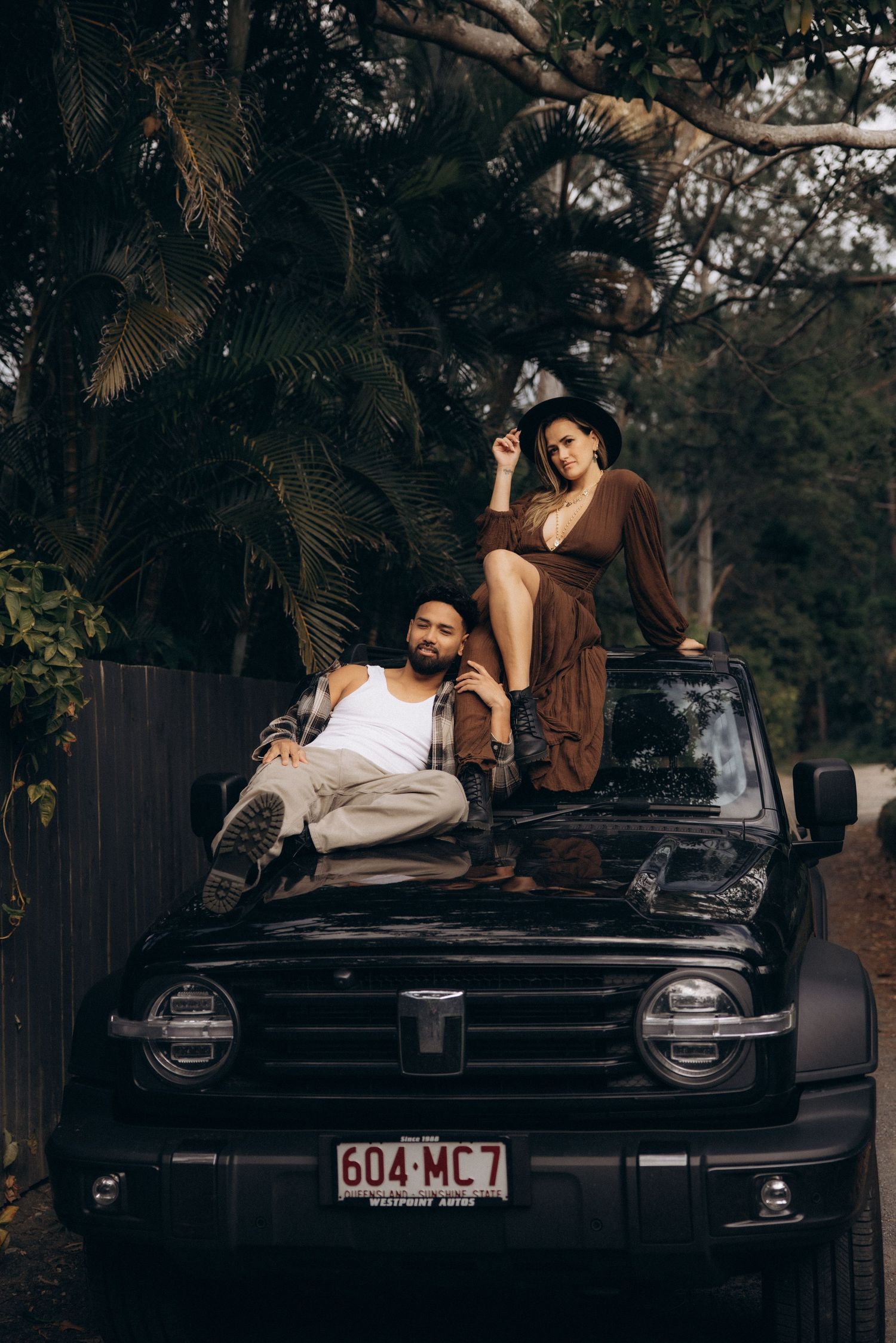 A stylish couple poses on a vintage black car during a photoshoot.