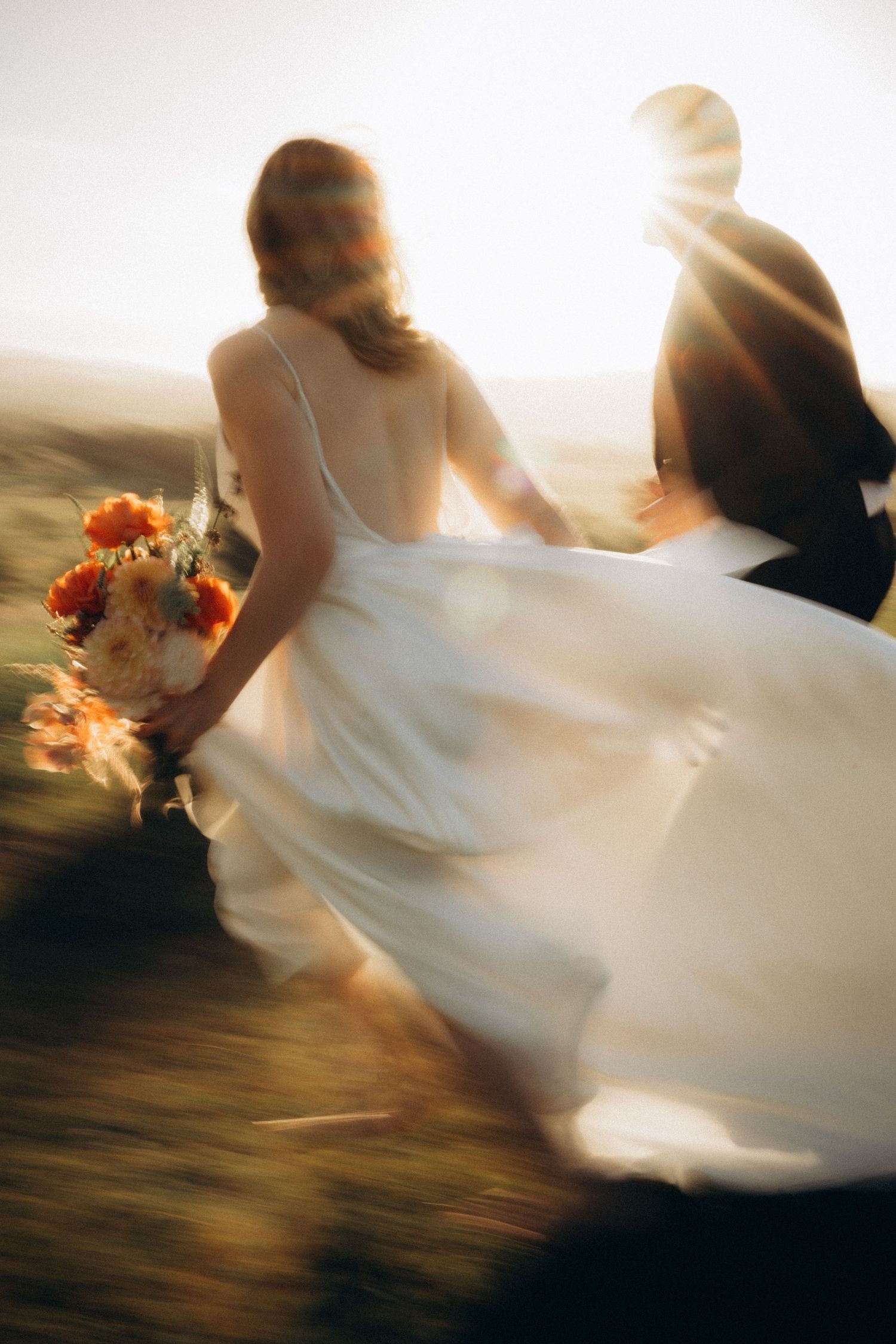 Bride in white gown holding orange bouquet runs with groom toward golden sunset, motion blur capturing joyful wedding moment.