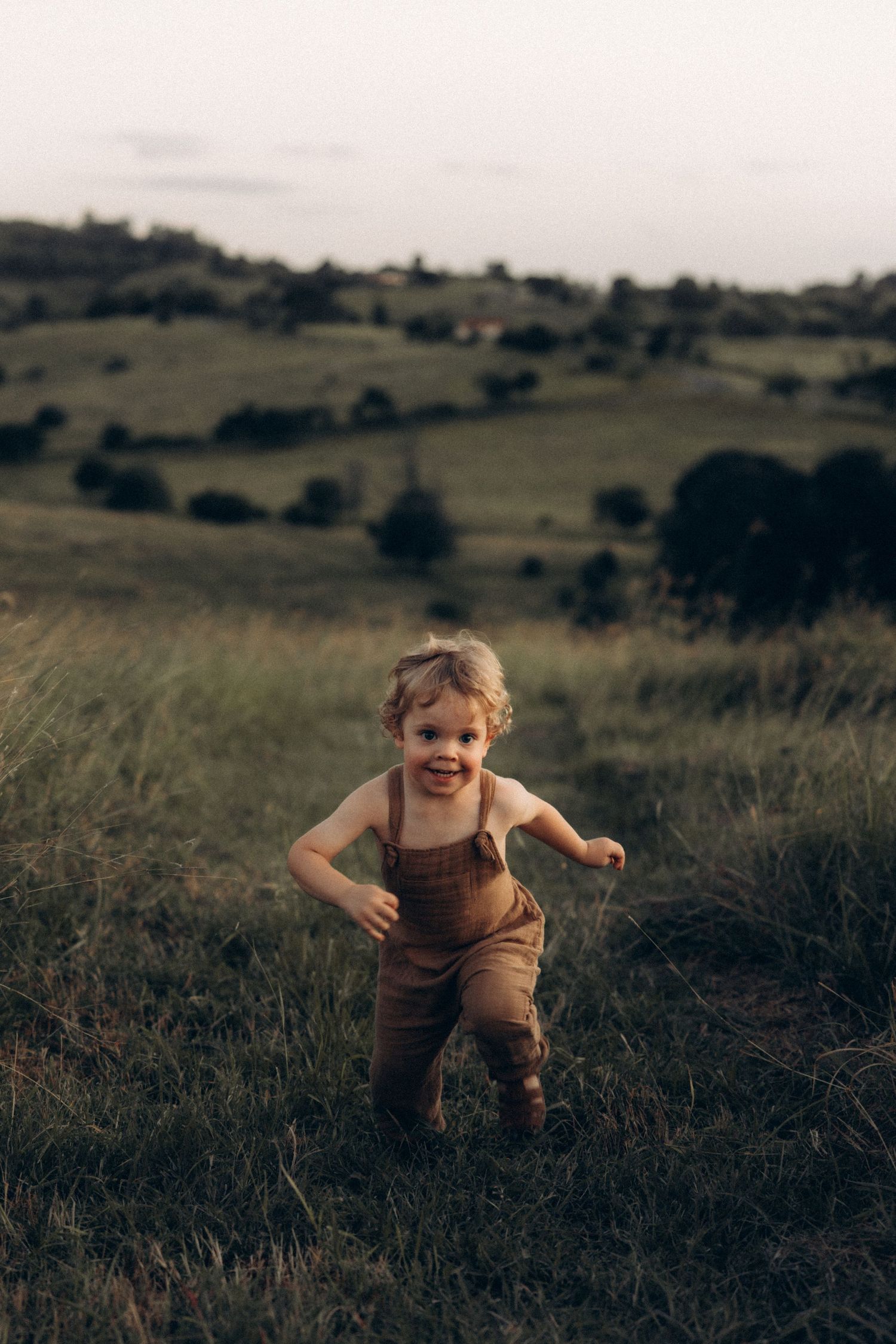 A child runs joyfully through tall grass in a rolling countryside landscape.