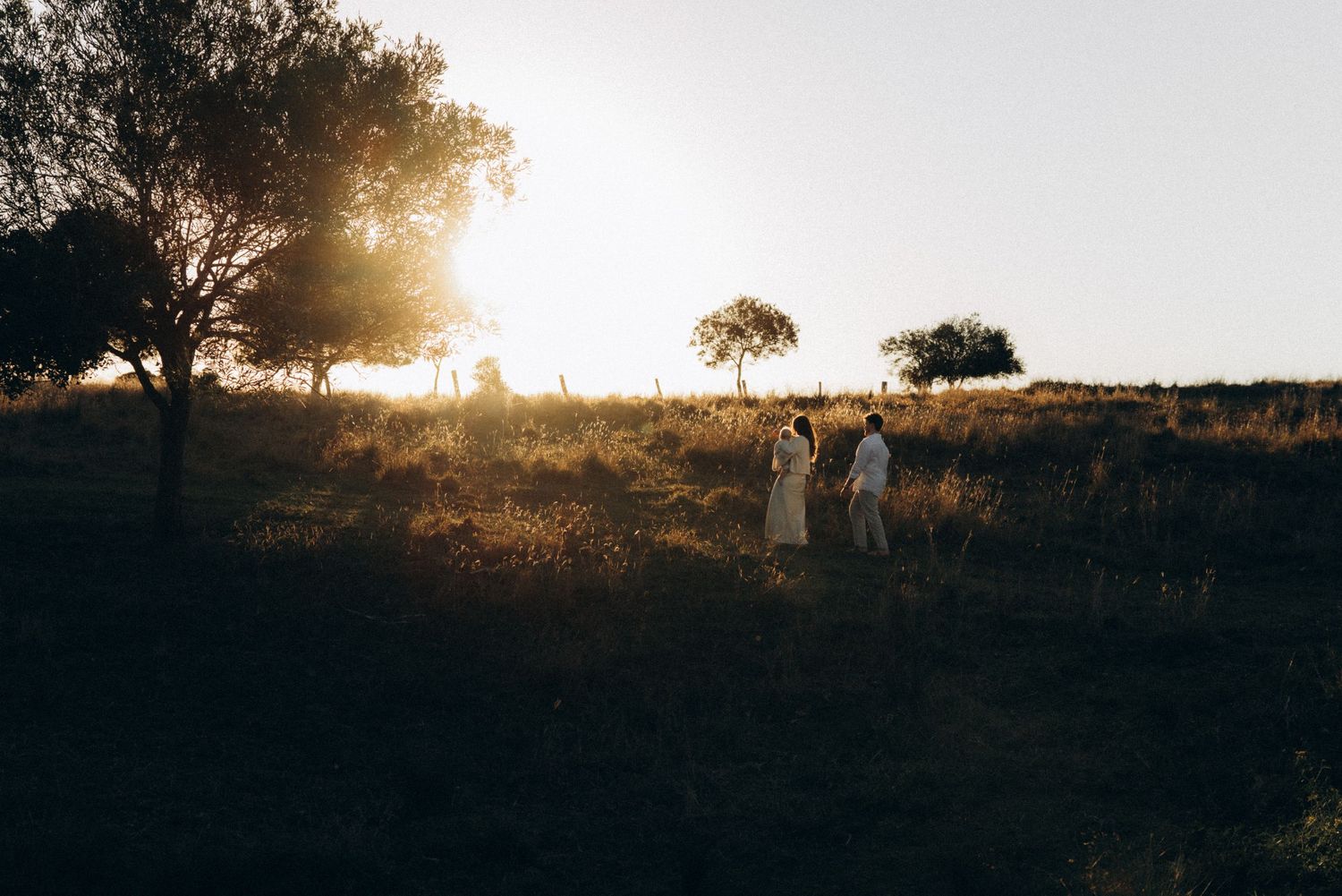 Two people walk through golden sunlit fields at sunset with trees silhouetted against the glowing horizon.