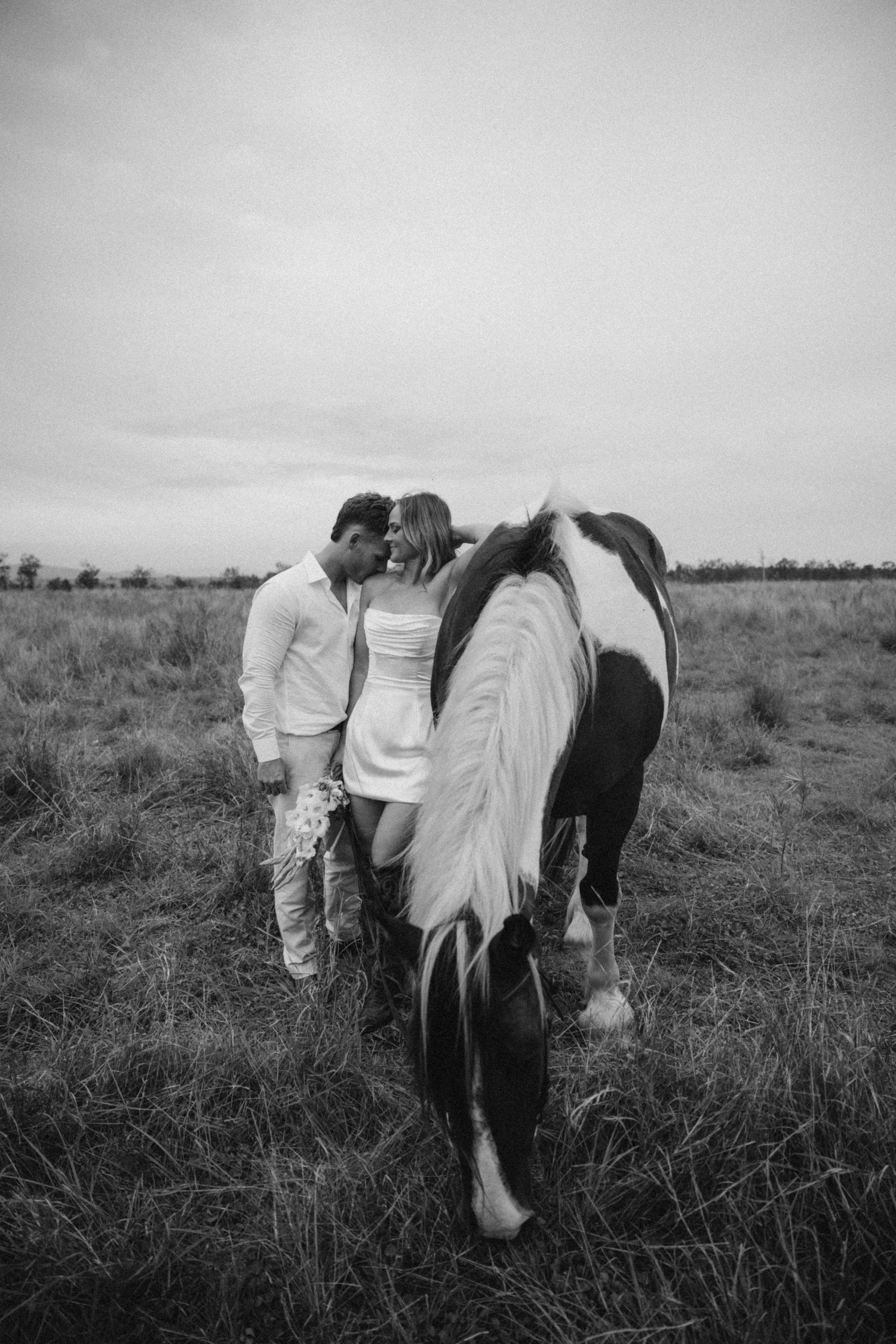 Black and white photo of a couple sharing an intimate moment with a horse in a field.