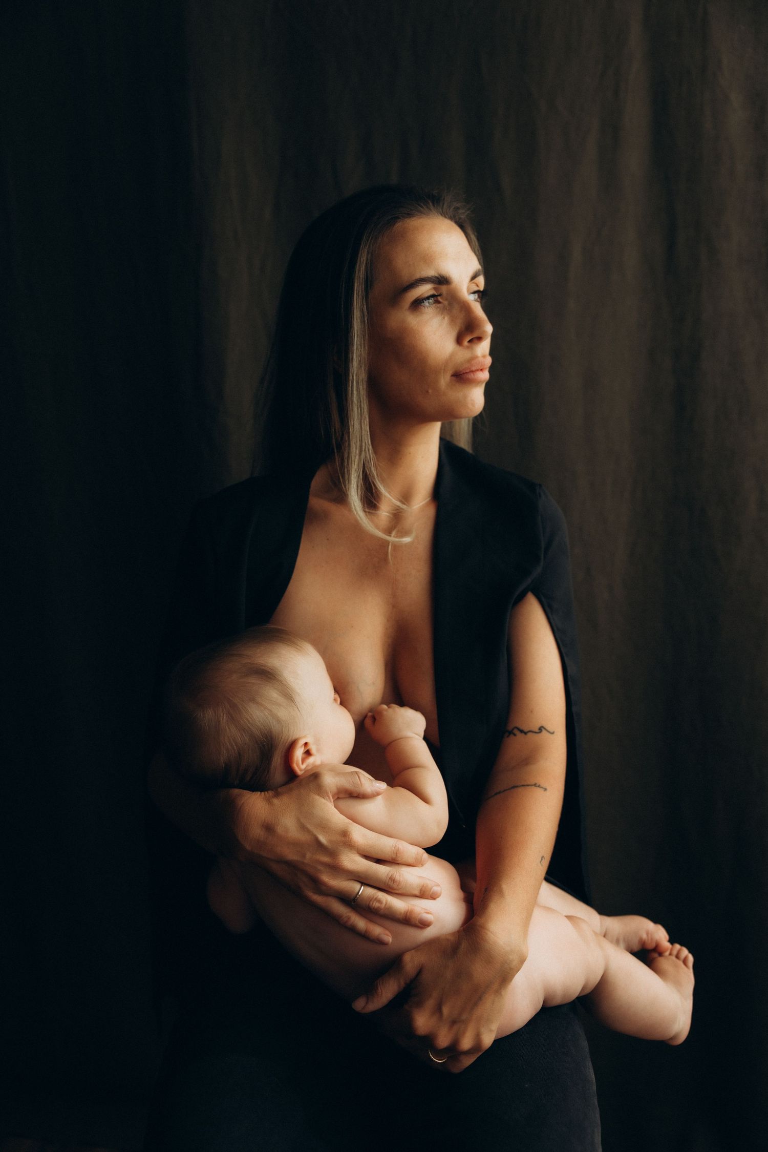 A mother breastfeeding her infant while gazing thoughtfully, set against a dark studio background.