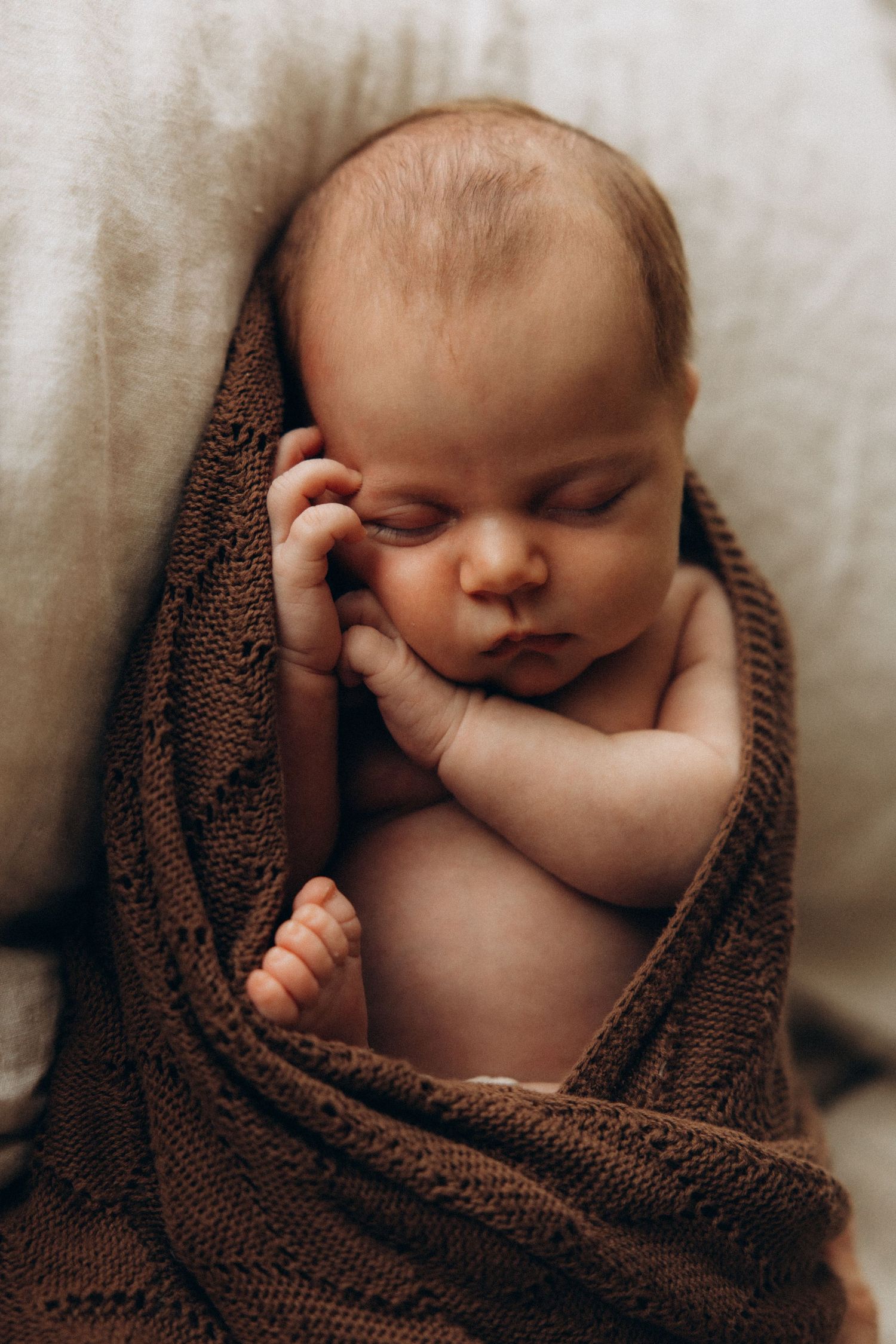 Sleeping newborn wrapped in brown knit blanket with tiny hand near face.