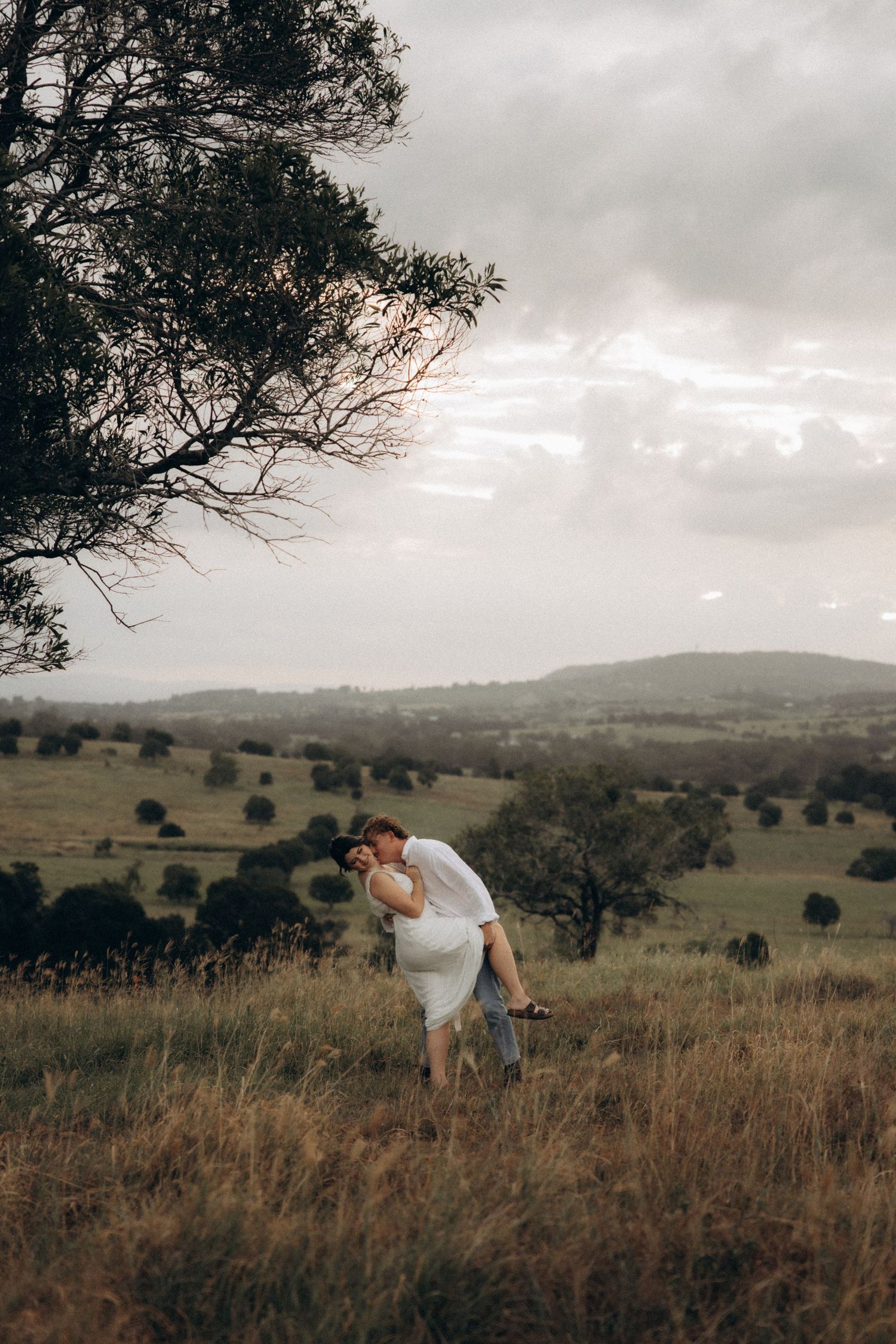 A couple dances together in a vast field with rolling hills and cloudy skies in the background.