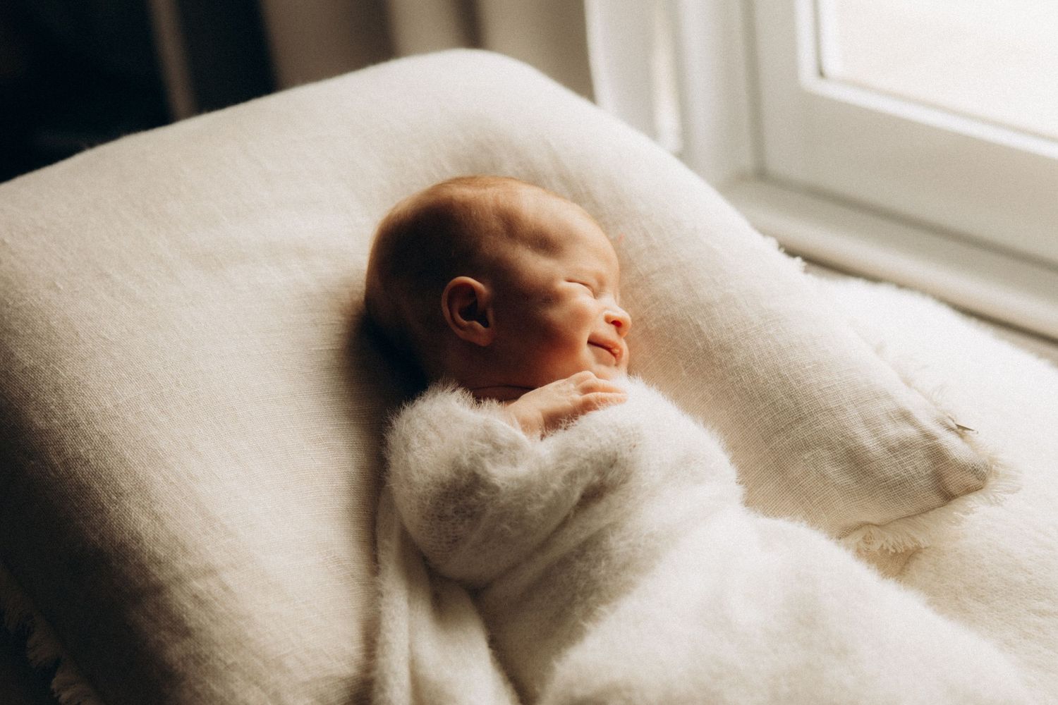 Sleeping newborn baby wrapped in soft white blanket by window.