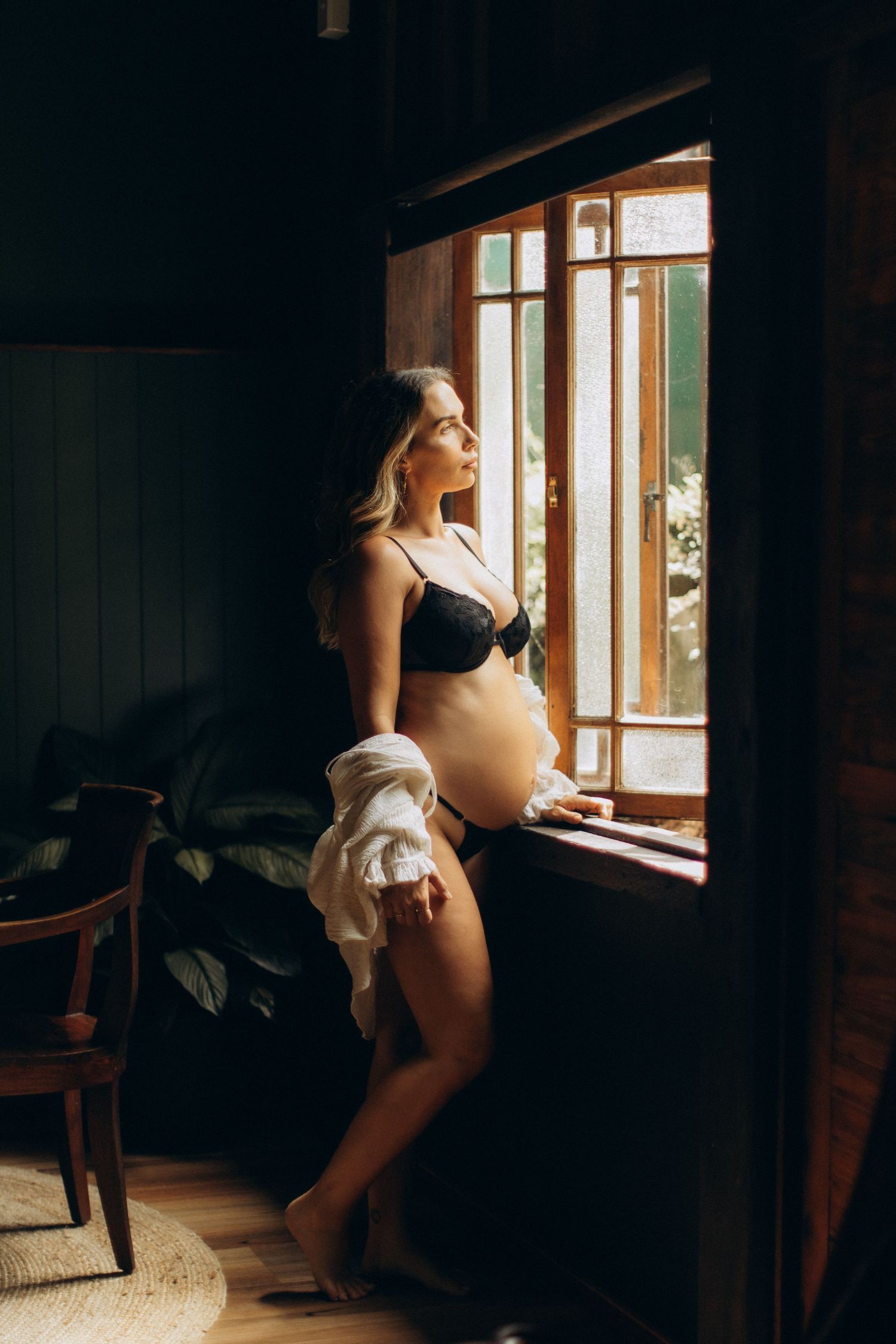 Dramatic window lighting illuminates a moody indoor boudoir photoshoot with dark shadows and natural light.