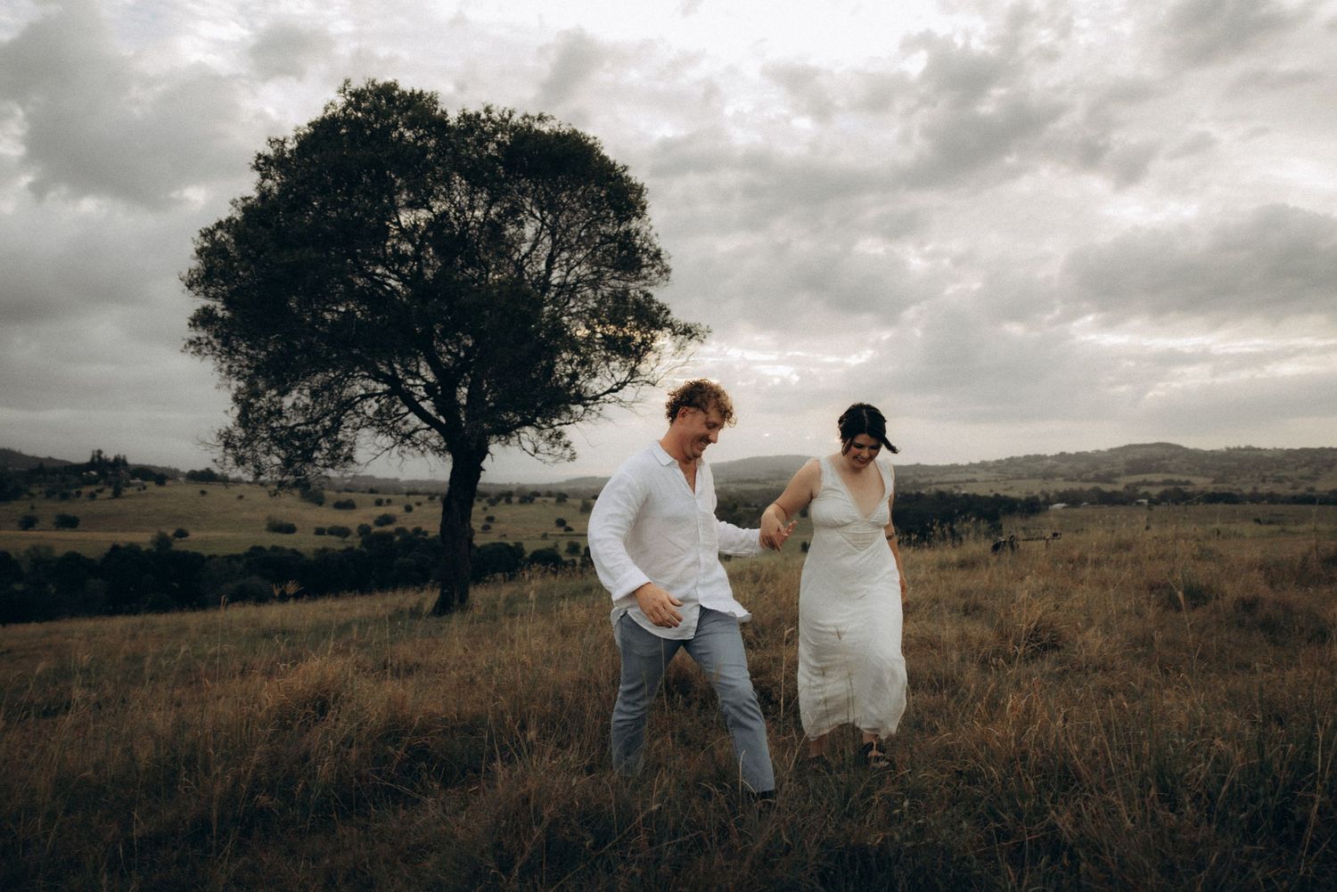 Two people share a playful moment together in a grassy field under cloudy skies.