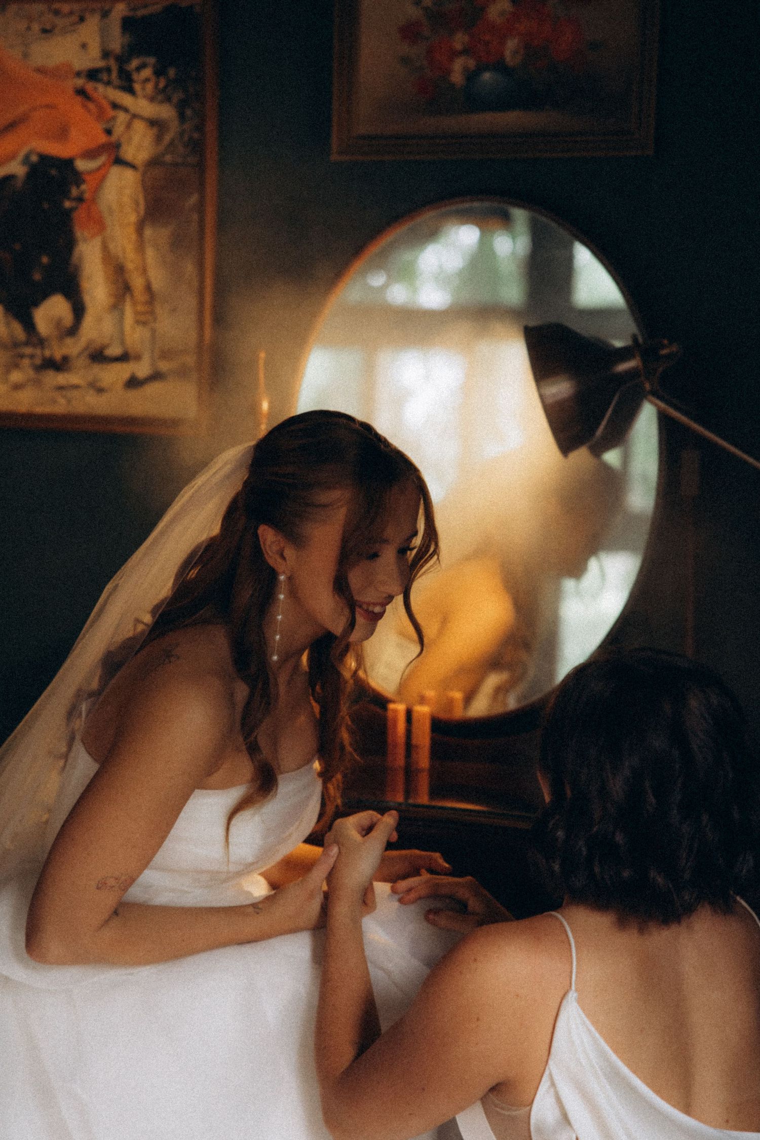 Bride in white gown and veil sits with bridesmaid in candlelit room with mirror and vintage artwork.