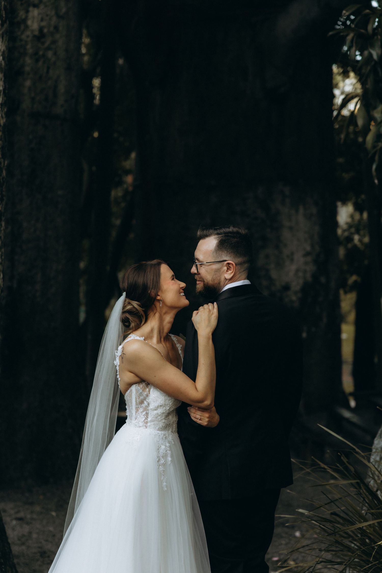 Bride and groom share a romantic moment among tall trees, her white gown and veil glowing in soft forest light.