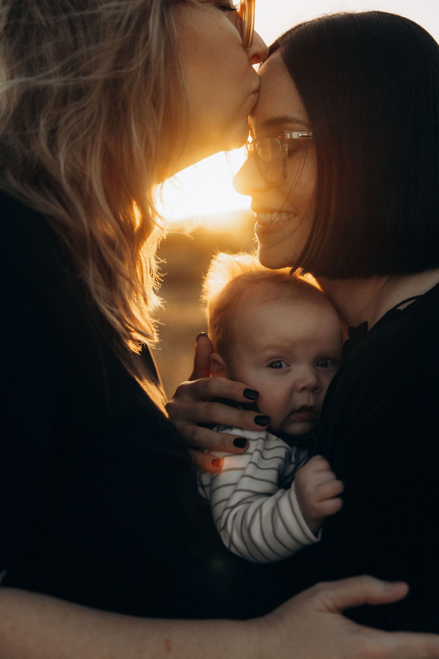 Two mothers sharing a kiss while holding their newborn baby in warm golden sunset light.