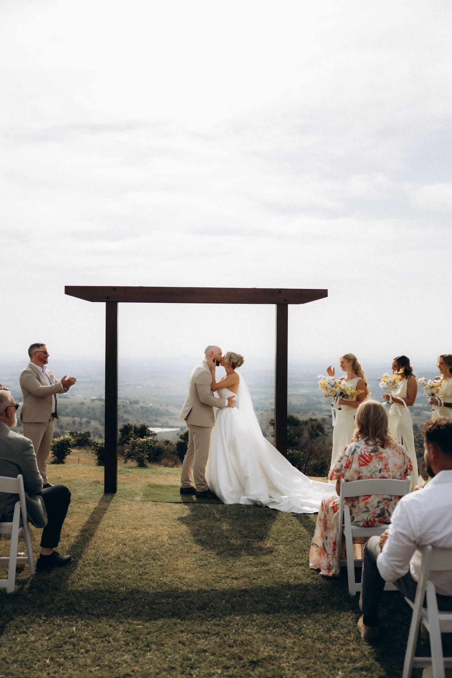 A wedding ceremony takes place on a hilltop with a wooden arch and scenic mountain backdrop.