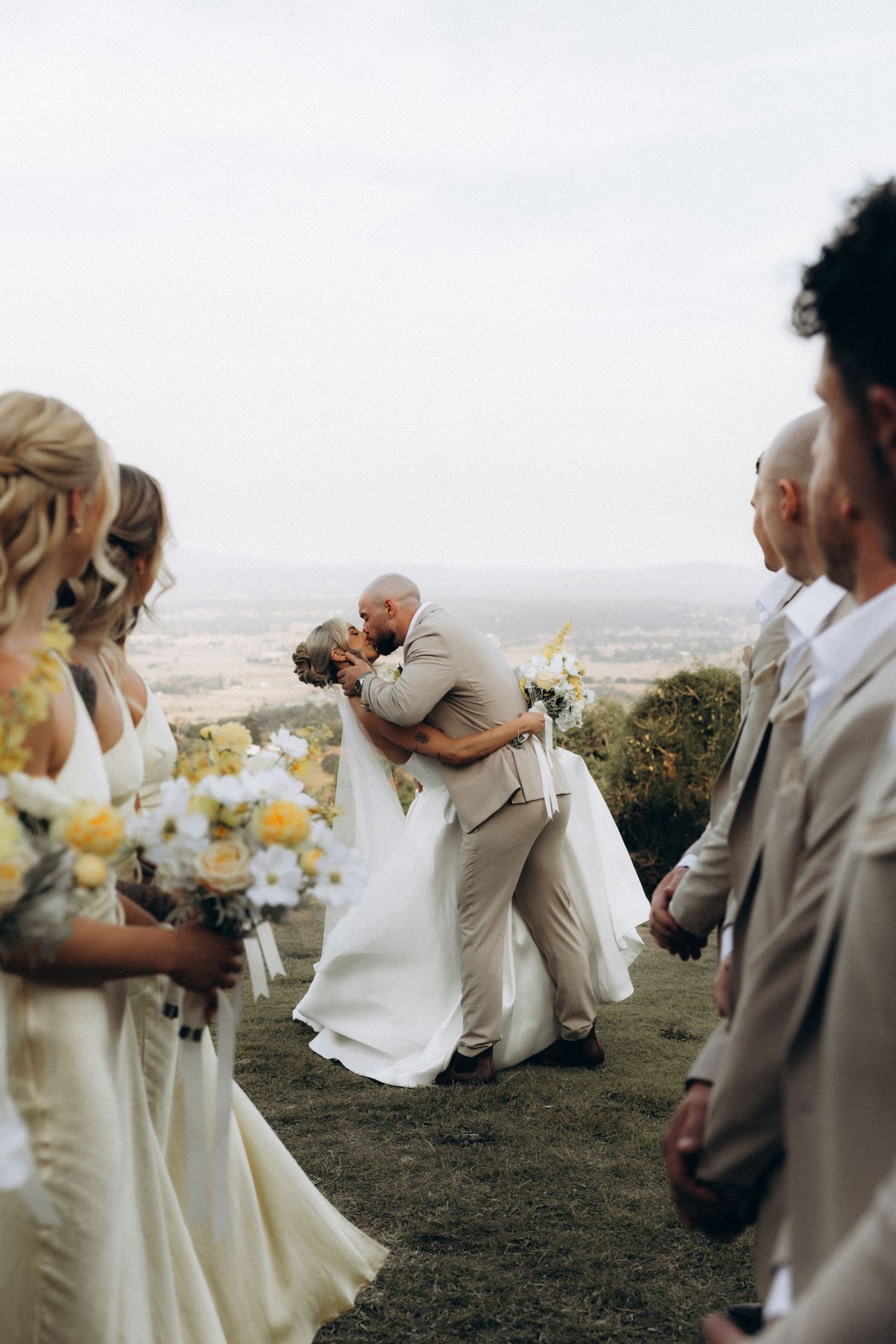 Wedding couple shares romantic dip kiss while wedding party looks on at scenic overlook.