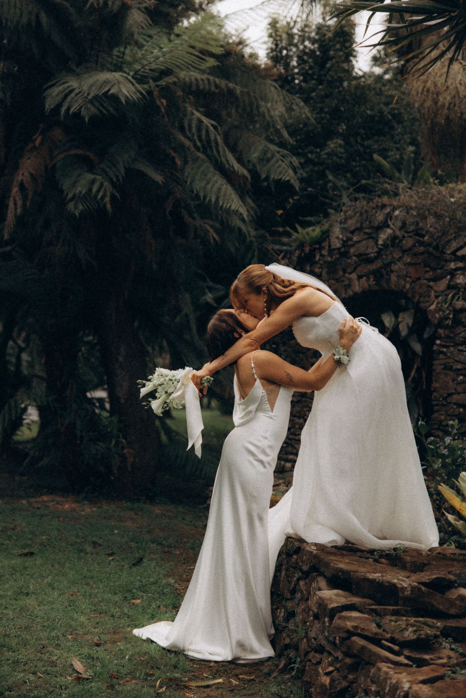 Two brides in white gowns share a romantic dip kiss surrounded by lush tropical greenery.