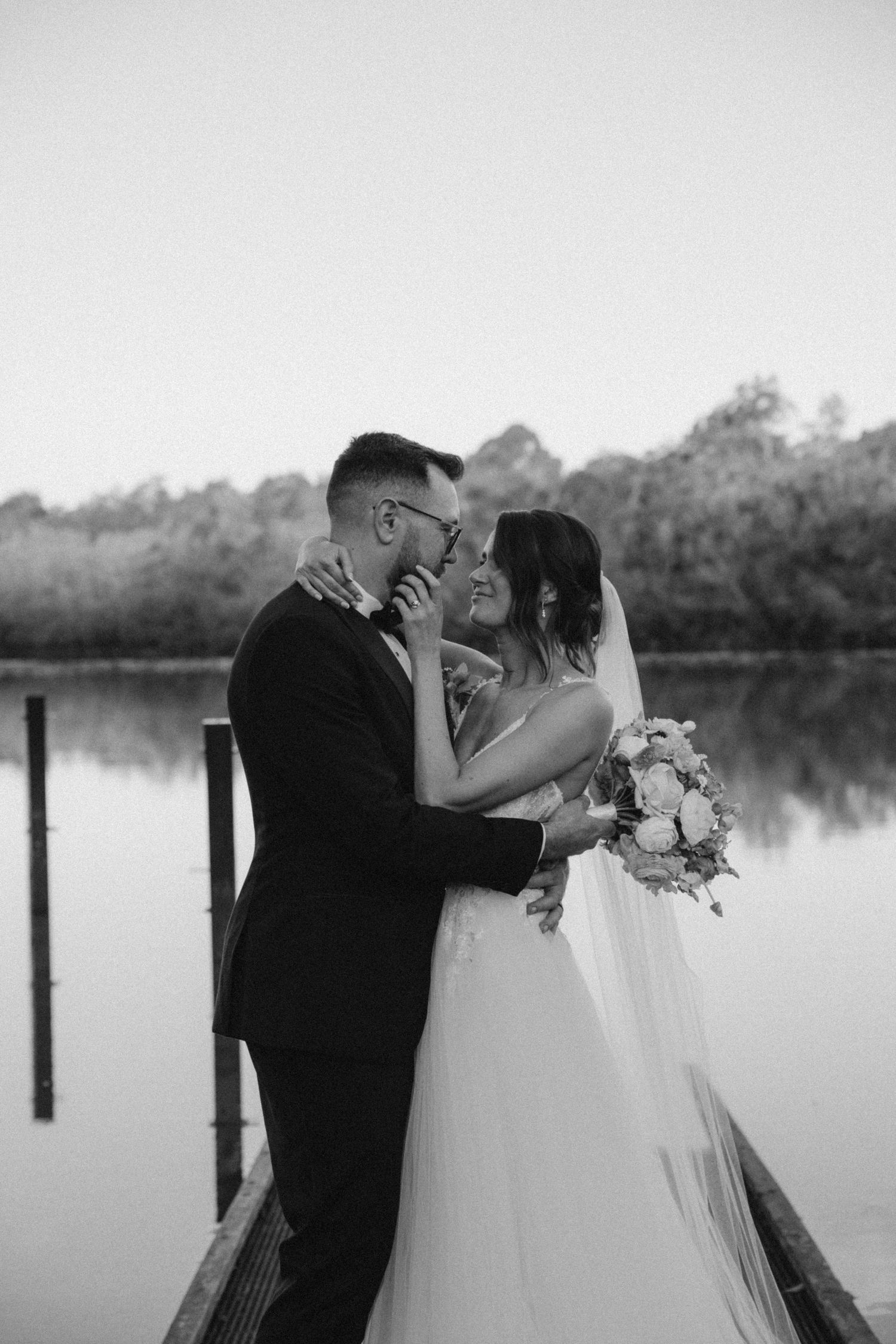 Black and white photo of a bride and groom sharing a romantic moment on a dock by a lake, bride holding a bouquet.