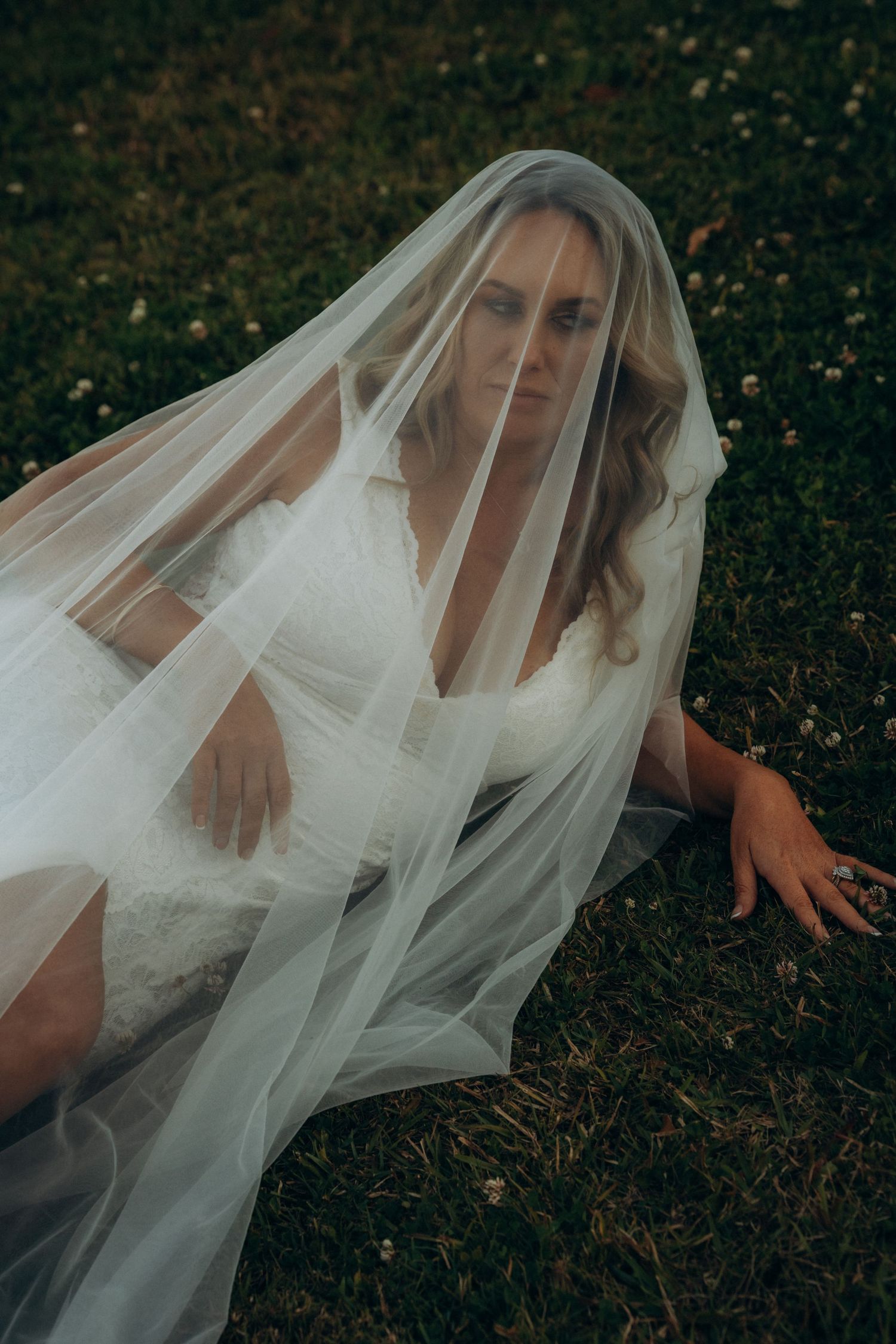 Bride in white lace wedding dress sitting on grass, draped with flowing sheer veil, outdoor bridal portrait photography.