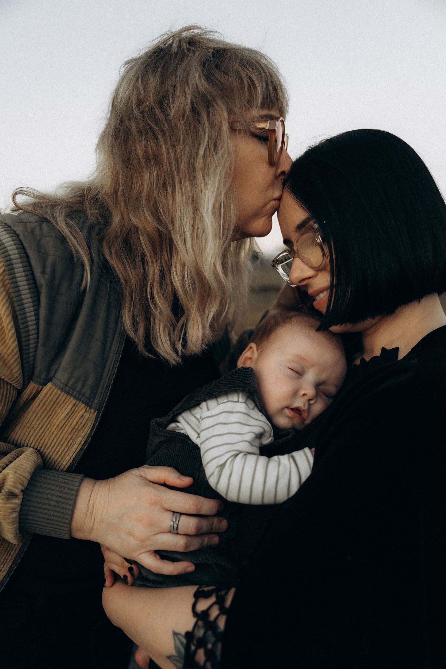 Two mothers sharing a tender forehead kiss while cradling their sleeping newborn baby together.