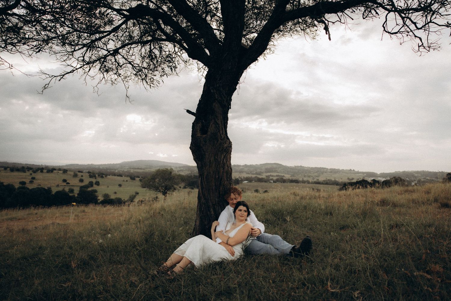 A romantic couple shares tender moments under a majestic tree in a rural landscape.