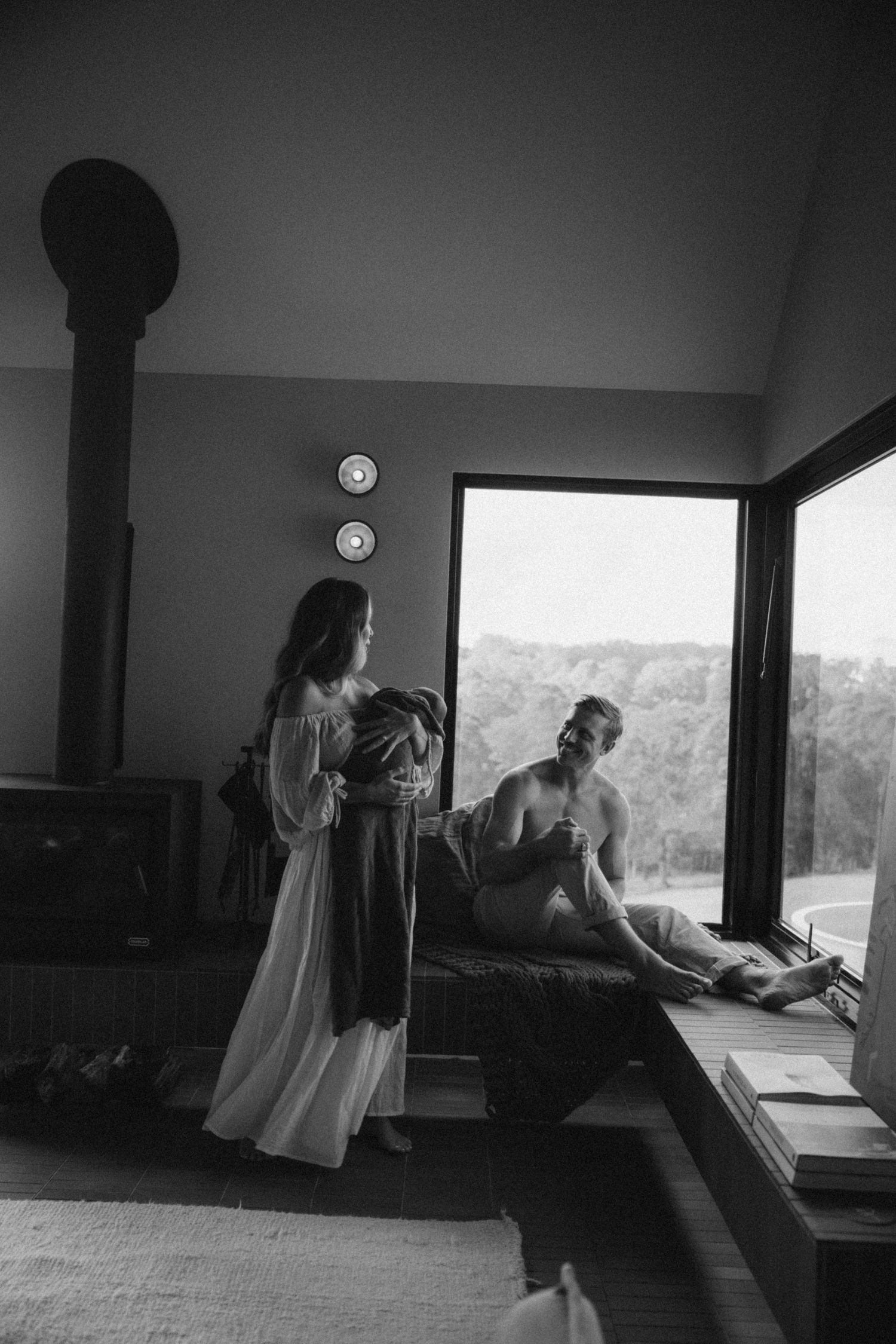 Couple with newborn baby standing by large window in modern rustic room.
