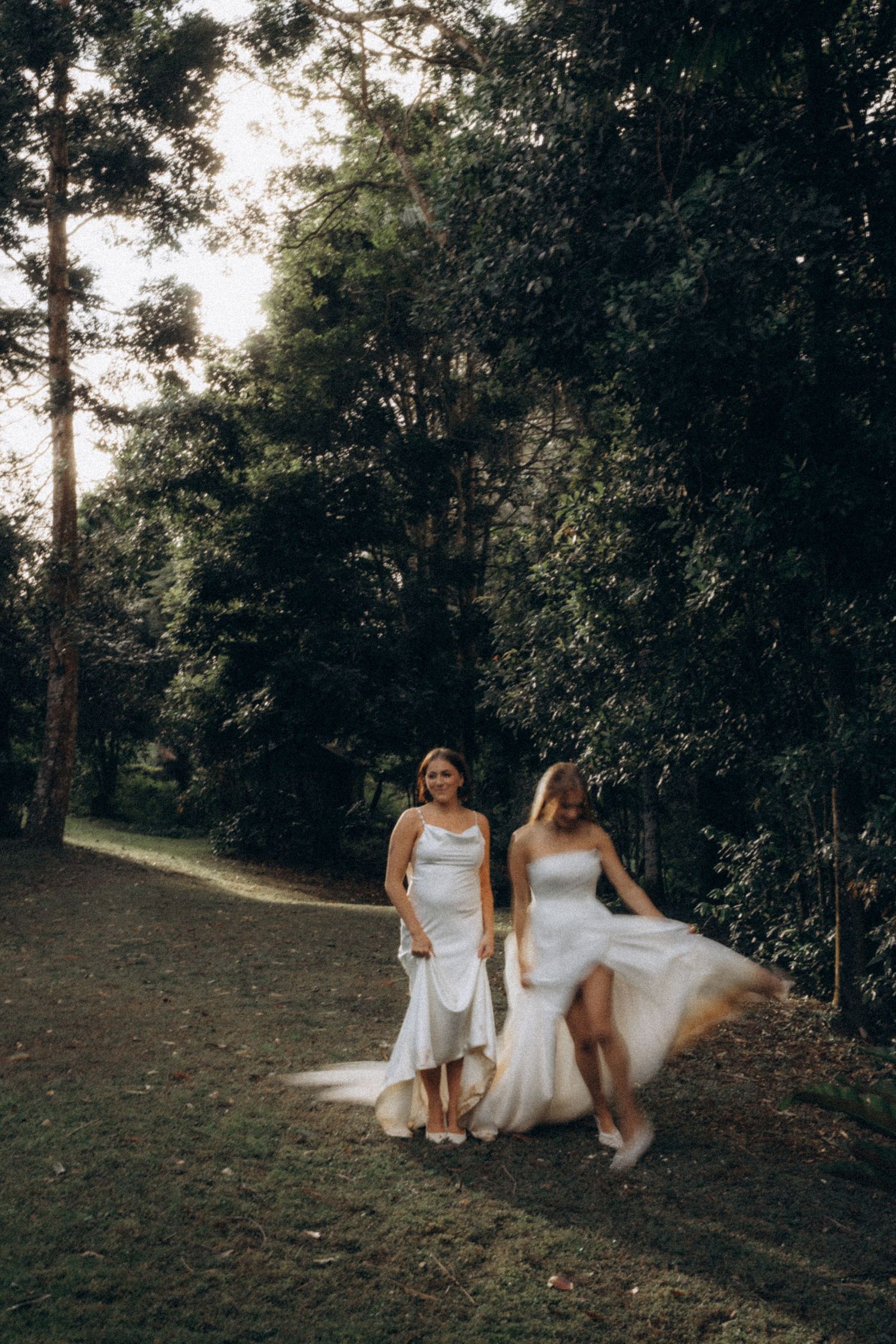 Two brides in white wedding dresses walk along a forest path surrounded by lush green trees.